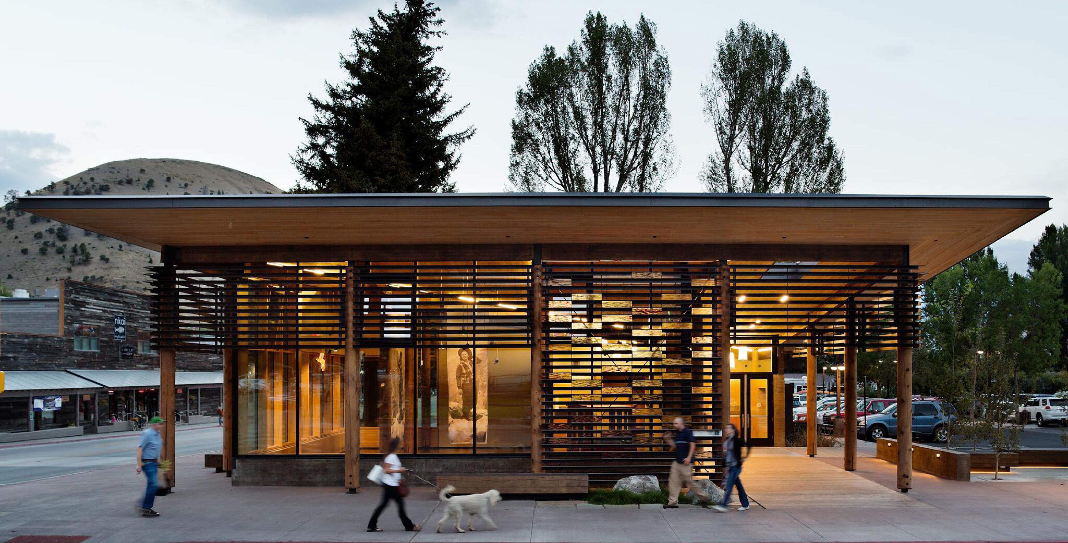 A contemporary building with a wide, flat wooden roof and an exterior facade of horizontal wood slats and large glass windows. The warmly lit interior contrasts with the cool light of dusk, as people walk on the sidewalk in front, with a street, older buildings, and a distant mountain under a cloudy sky in the background.