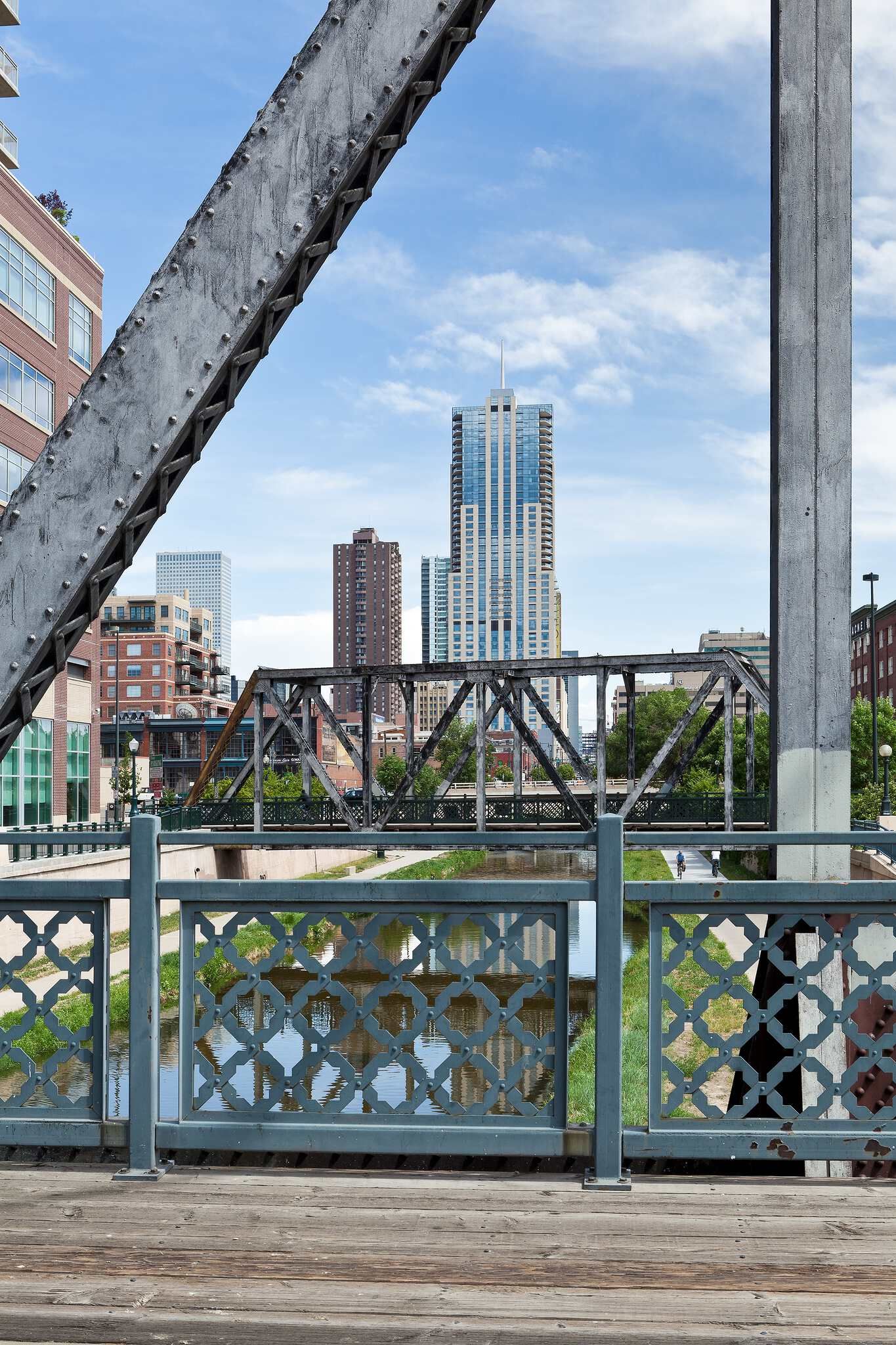 A city view framed by a large diagonal metal truss and a concrete pillar in the foreground. Below, the bridge's wooden deck and an ornate gray metal railing overlook a river. A green path with a distant cyclist leads to an old metal truss bridge spanning the water. In the background, a modern skyscraper with a spire rises above other city buildings under a bright blue sky with white clouds.