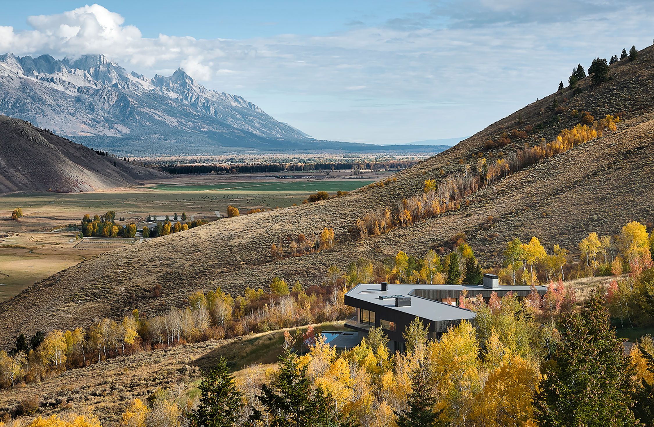 Aerial landscape view of a modern boomerang-shaped home nestled in a valley with autumn foliage and mountain ranges in the background.
