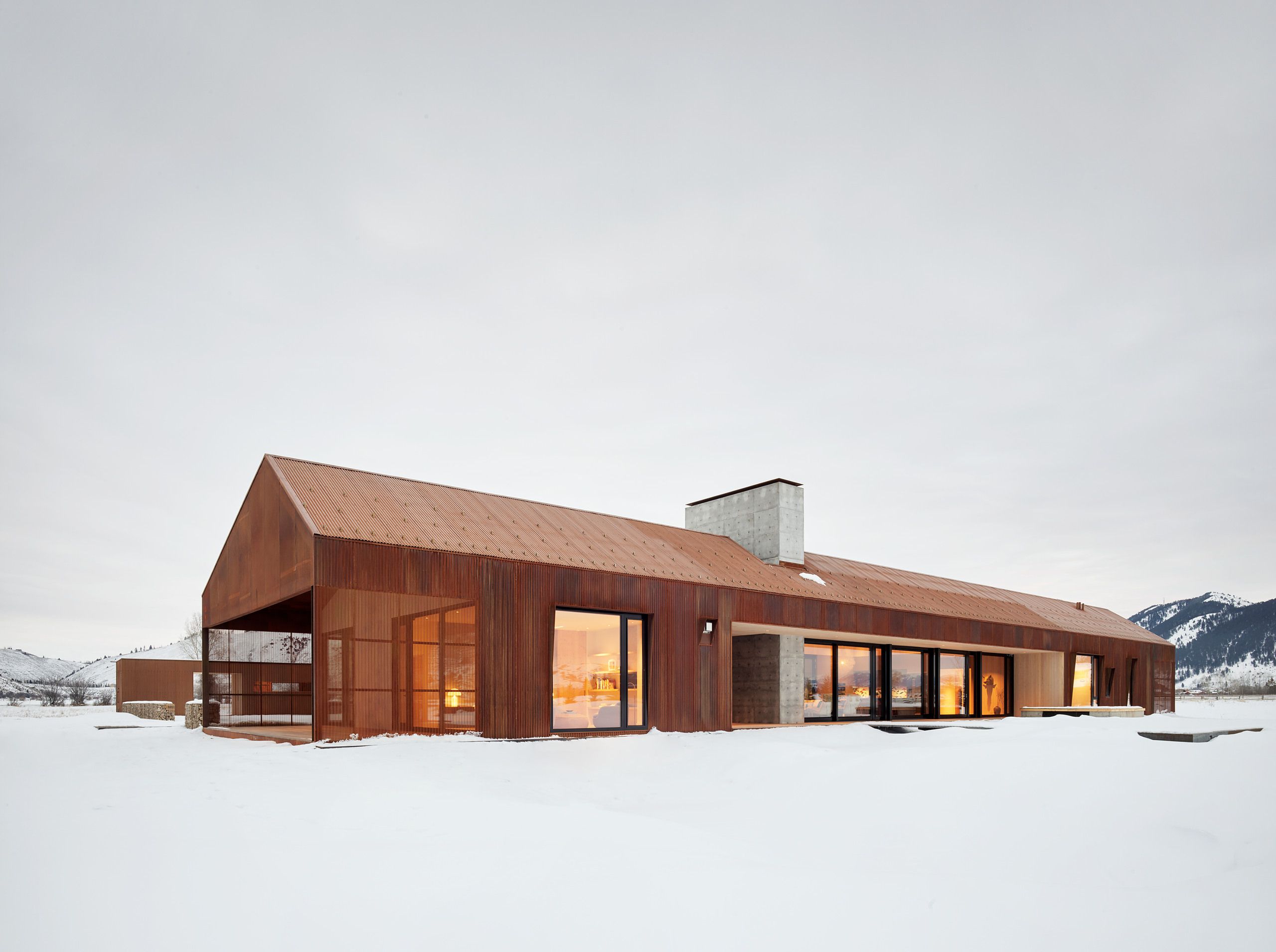A modern, linear home crafted from rust-colored Corten steel and concrete, featuring a gabled roof and large windows with warm interior lighting. The house is set in a vast, snow-covered winter landscape with distant, tree-lined mountains under an overcast sky.