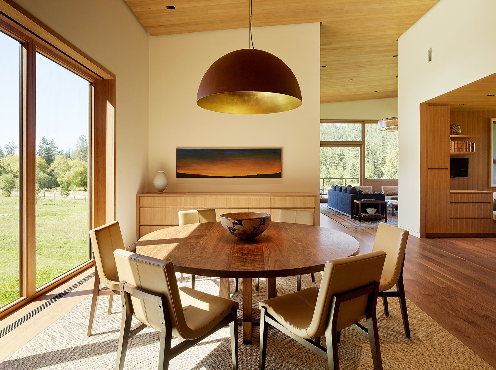 Modern open-concept dining room with a round dark wood table, six tan leather chairs, and a large bronze pendant light with a gold interior. Floor-to-ceiling windows reveal a green outdoor landscape. A light wood credenza with horizontal sunset artwork and a vase sits against the wall, with an adjacent living area visible.