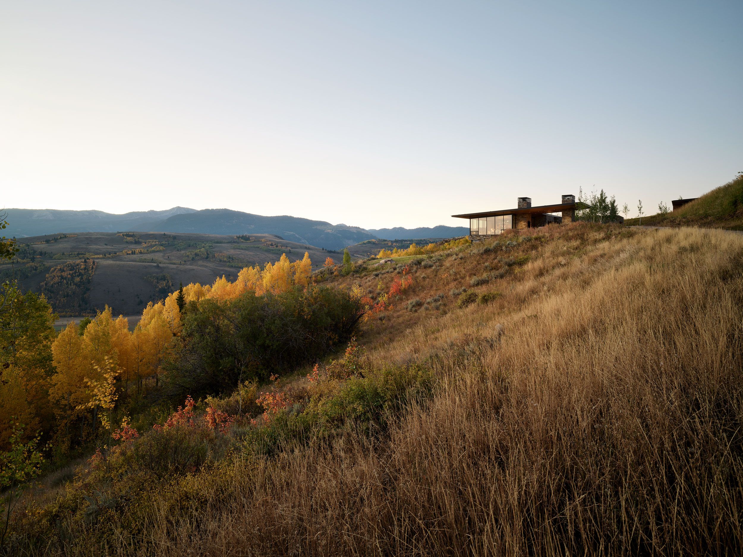 Modern mountain home with expansive glass windows and natural stone chimneys, perched on a golden hillside of dry grass. It overlooks a valley vibrant with yellow and orange autumn foliage and distant rolling mountains under a soft, pale sky.