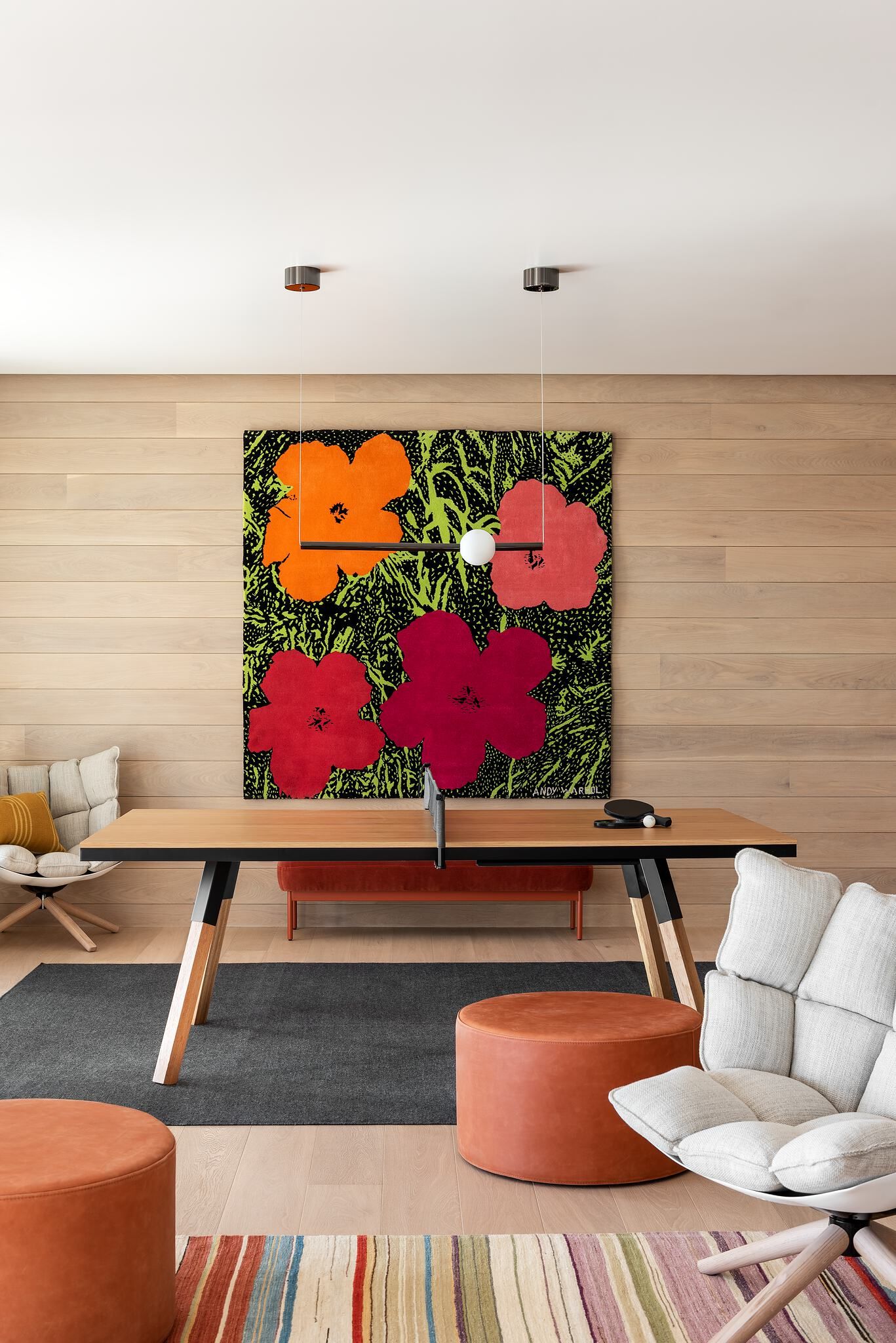 Modern game room featuring a wooden ping-pong table with black accents, centered in front of a large Andy Warhol Flowers pop art print with vibrant orange and red blossoms on a dark green and black background. The room has light wood-paneled walls and floor, a dark grey area rug, two cream upholstered armchairs, and two round orange leather ottomans, all illuminated by modern black and white pendant lights.