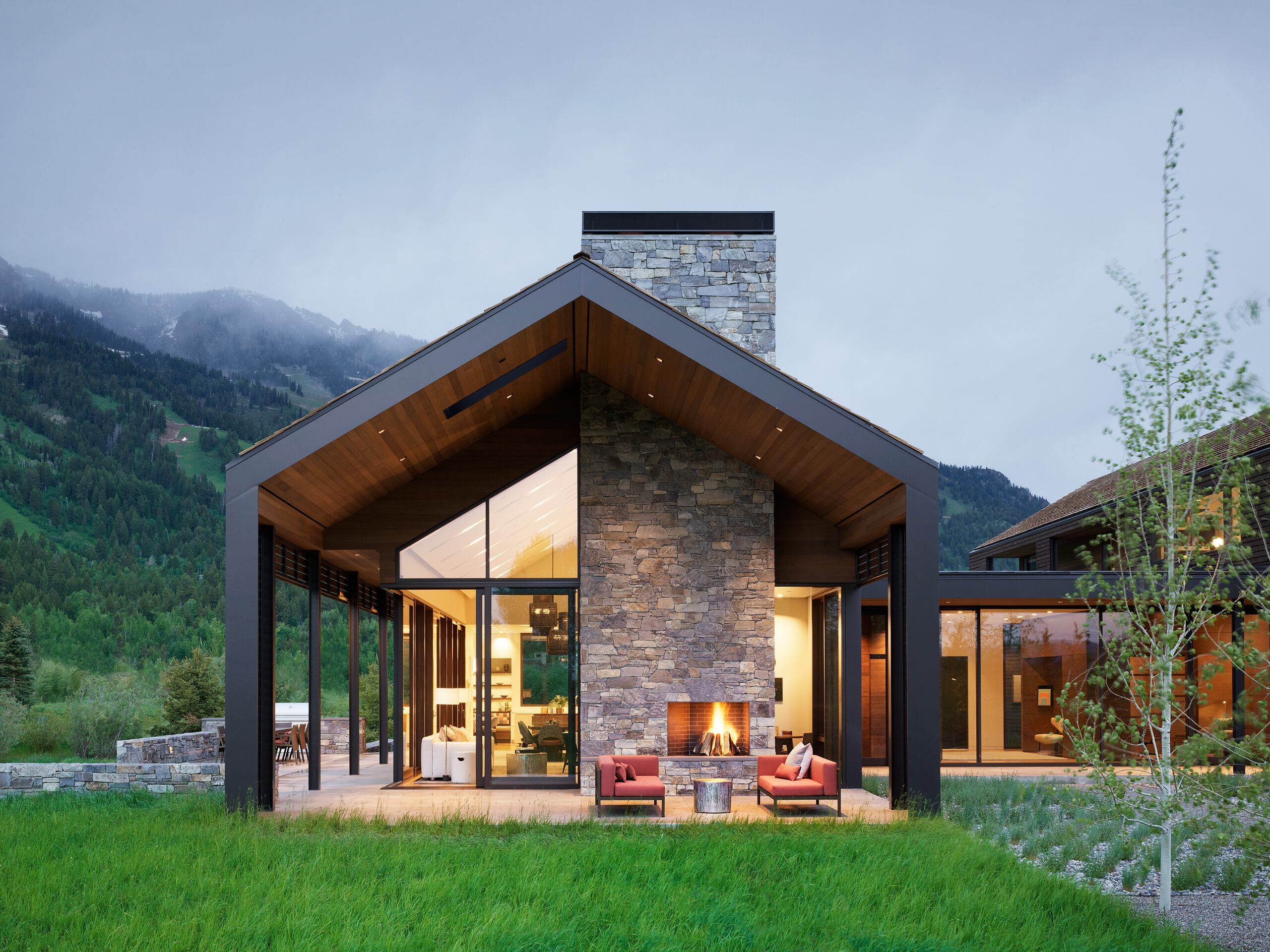Contemporary mountain home with a stone and floor-to-ceiling glass exterior, featuring a covered patio with an outdoor fireplace and lounge seating. Set against a backdrop of lush green mountains at dusk.