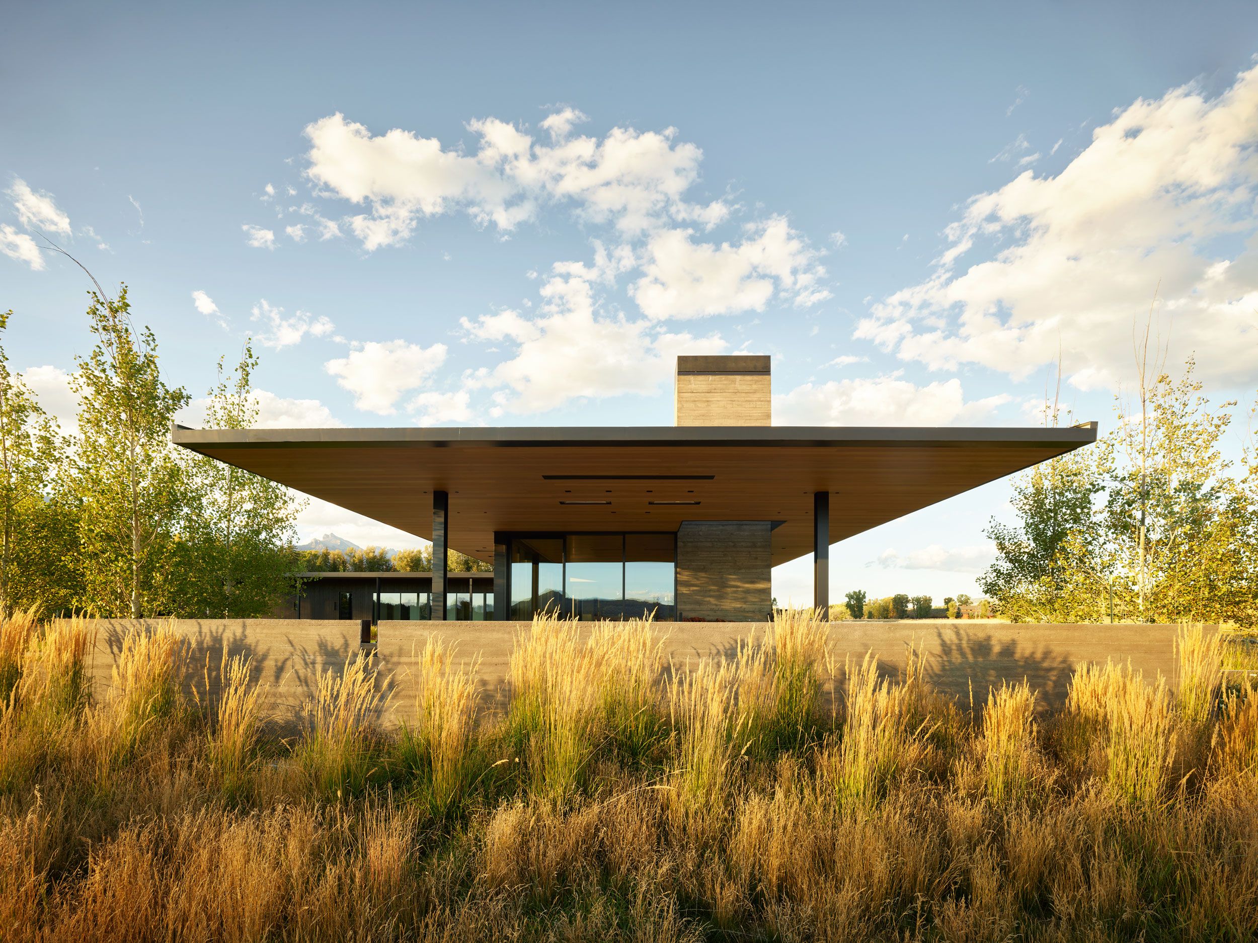 Modernist house with an expansive flat roof, concrete facade, and large glass windows, situated behind a low concrete wall and a vibrant field of golden prairie grass under a bright blue sky.