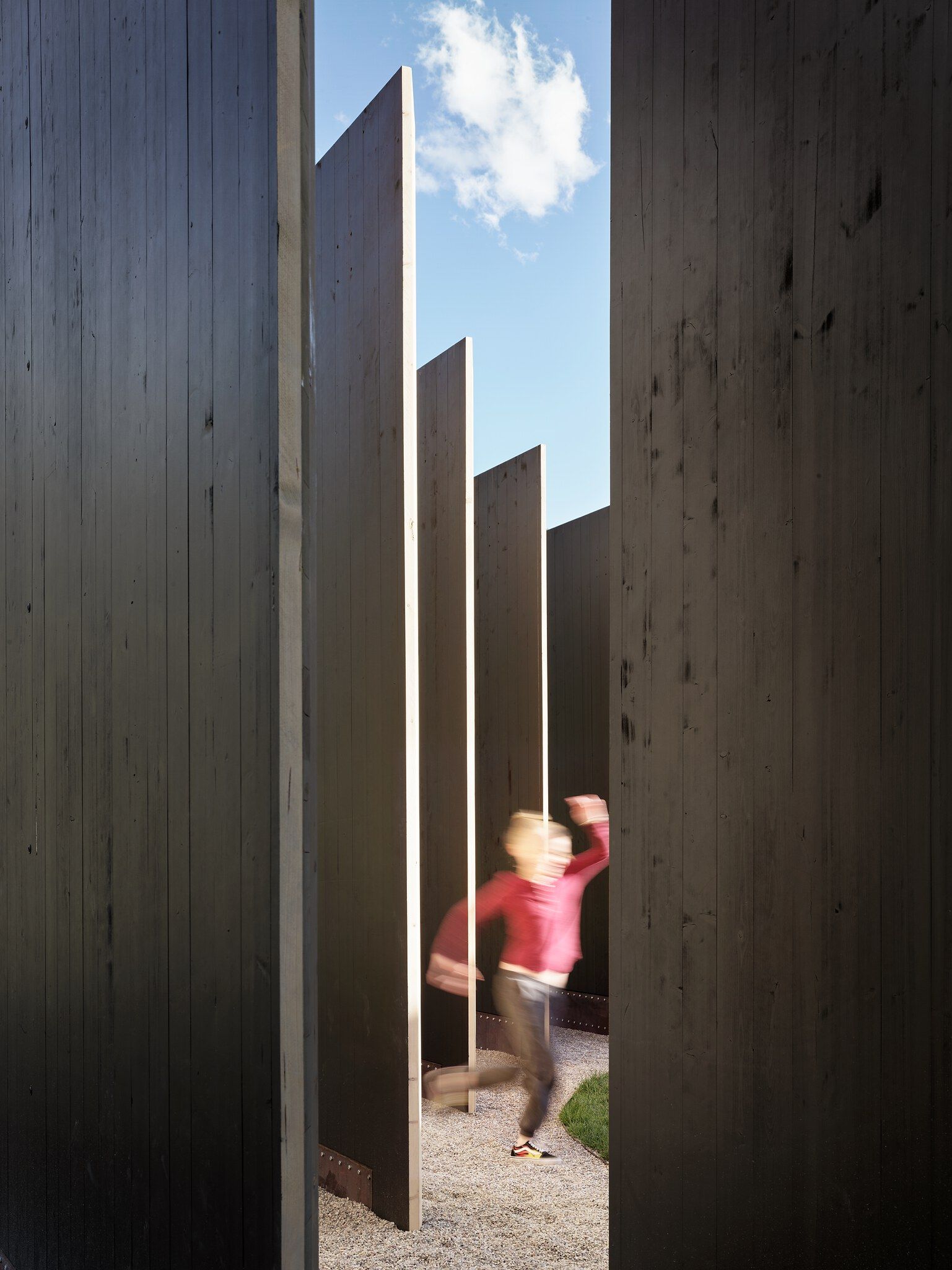 Blurred child running on a gravel path between tall, dark wooden architectural panels. Sunlight highlights the vertical edges of inner, lighter wooden walls, with glimpses of a blue sky and white cloud visible above.