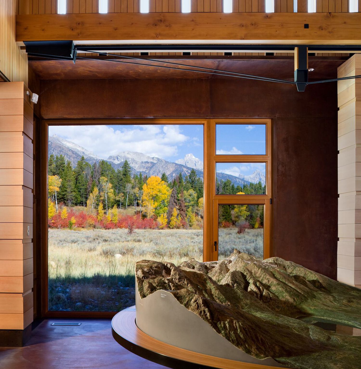 Warm wooden interior of a modern visitor center, featuring a slatted ceiling with vertical light fixtures and exposed beams. A large window frames a vibrant autumn landscape with golden and red trees, a grassy meadow, and rugged snow-capped mountains. In the foreground, a detailed topographic relief map of a mountain range rests on a curved display table.