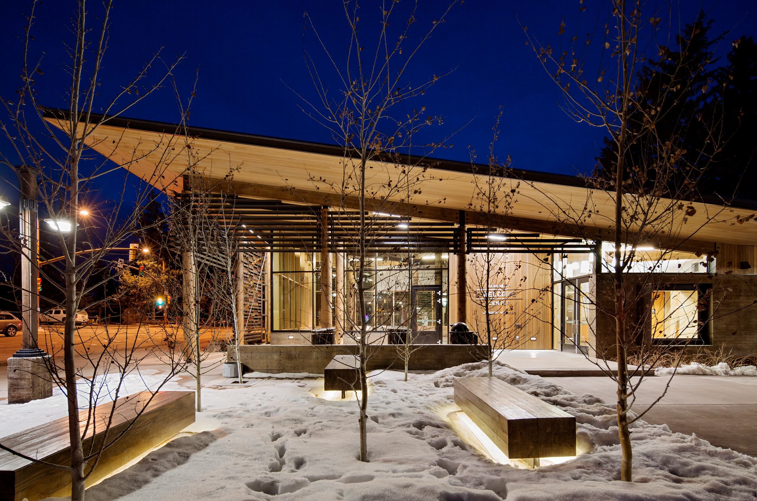 Modern welcome center building at dusk with a slanted wooden roof, glass facade, and warm interior lights, surrounded by snow-covered ground, bare trees, and illuminated wooden benches.