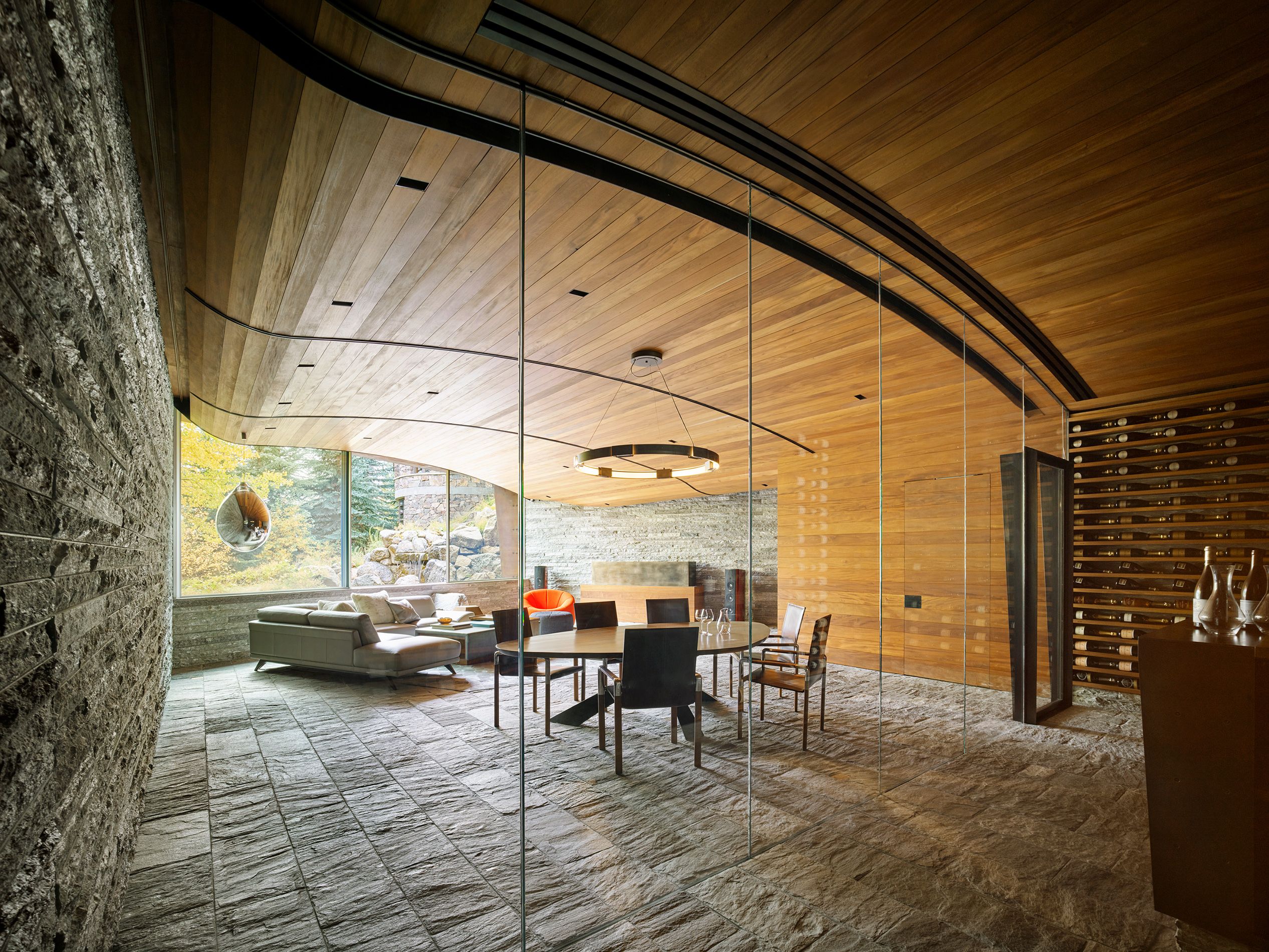 Glass-enclosed modern dining room adjacent to a wine display, featuring a wood ceiling and stone floors leading into a living area.