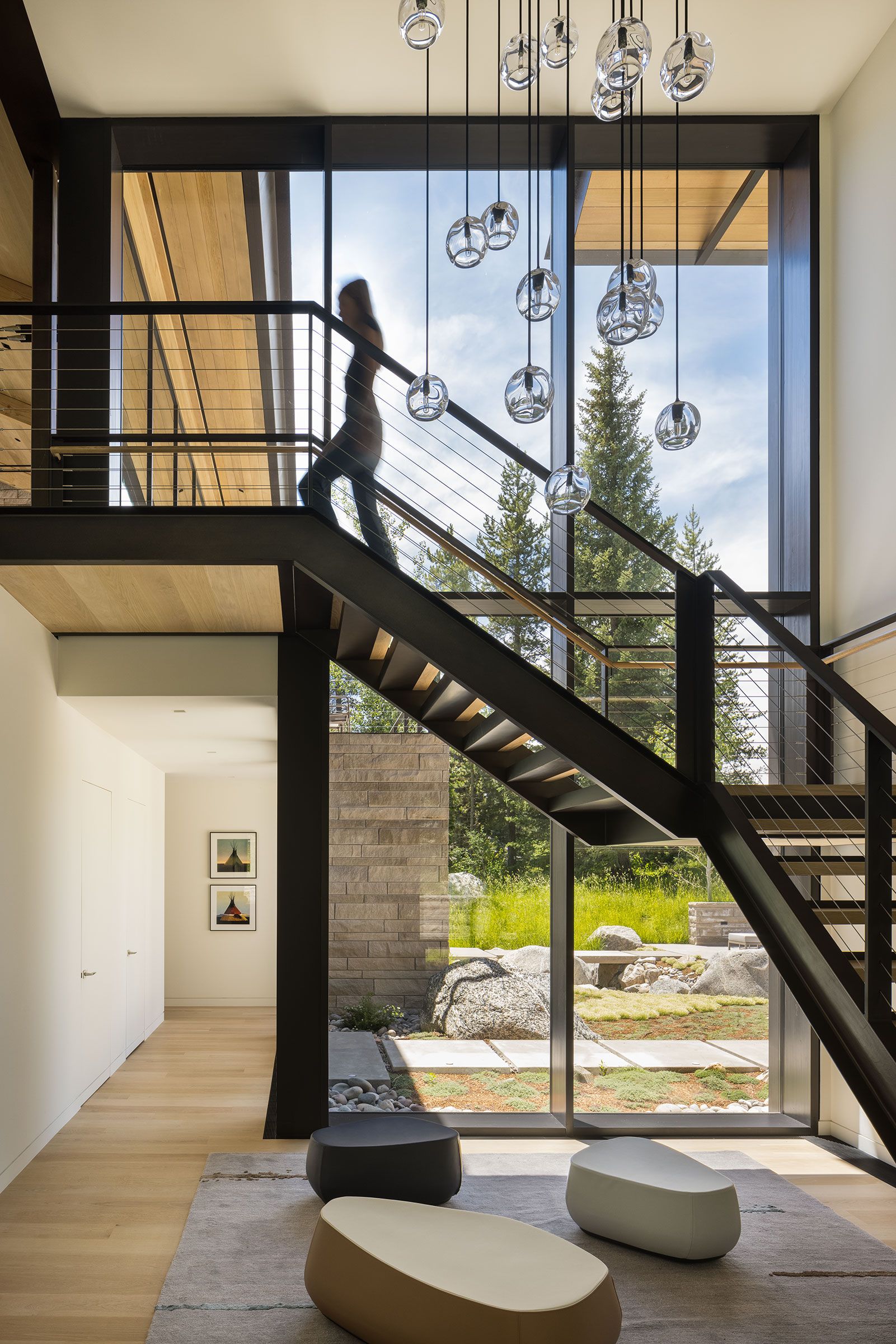 Interior staircase with a black steel frame and wire railings in a modern home, illuminated by a large multi-bulb glass pendant light fixture against a massive floor-to-ceiling window.