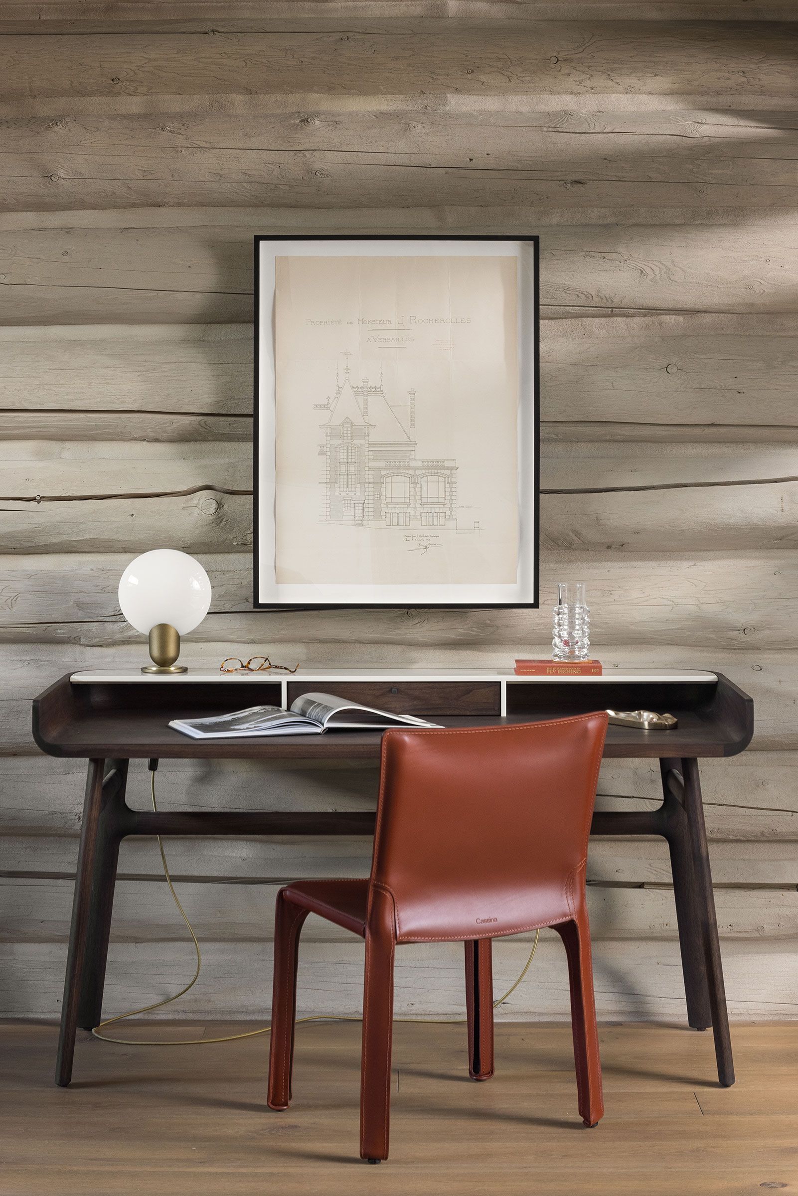 A dark wood writing desk paired with a brown leather chair against a rustic log wall, featuring a framed architectural drawing and a glowing globe lamp.