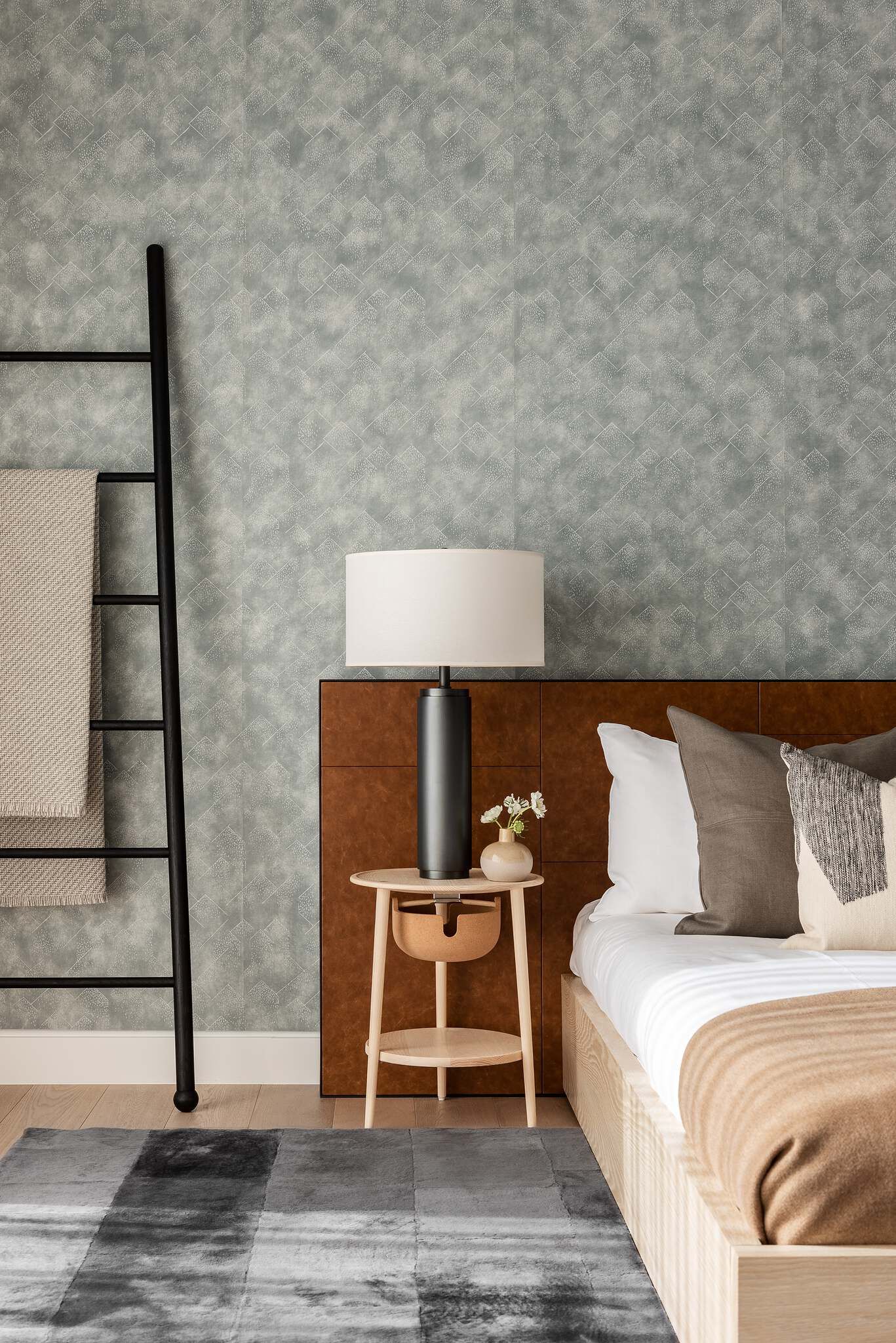 Modern bedroom interior with a light wood bed, brown leather headboard, and neutral bedding. A black ladder blanket rack stands next to blue-grey textured wallpaper featuring a subtle geometric pattern. A wooden nightstand holds a black lamp and small vase, with a grey striped rug on the floor.