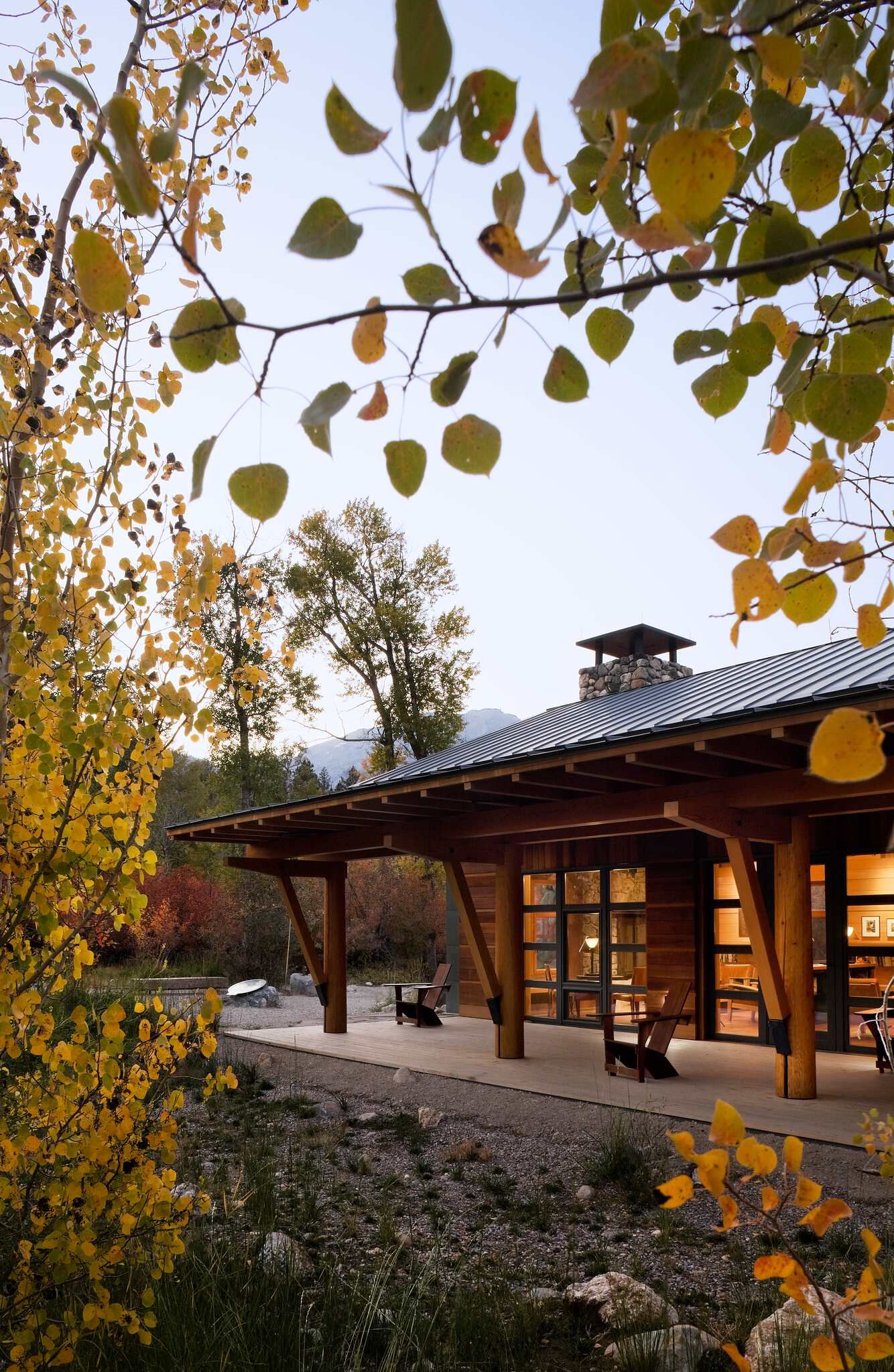 Modern rustic wooden home exterior at dusk, with a wide covered porch, large windows, and a metal roof, framed by vibrant golden aspen trees and distant fall foliage.