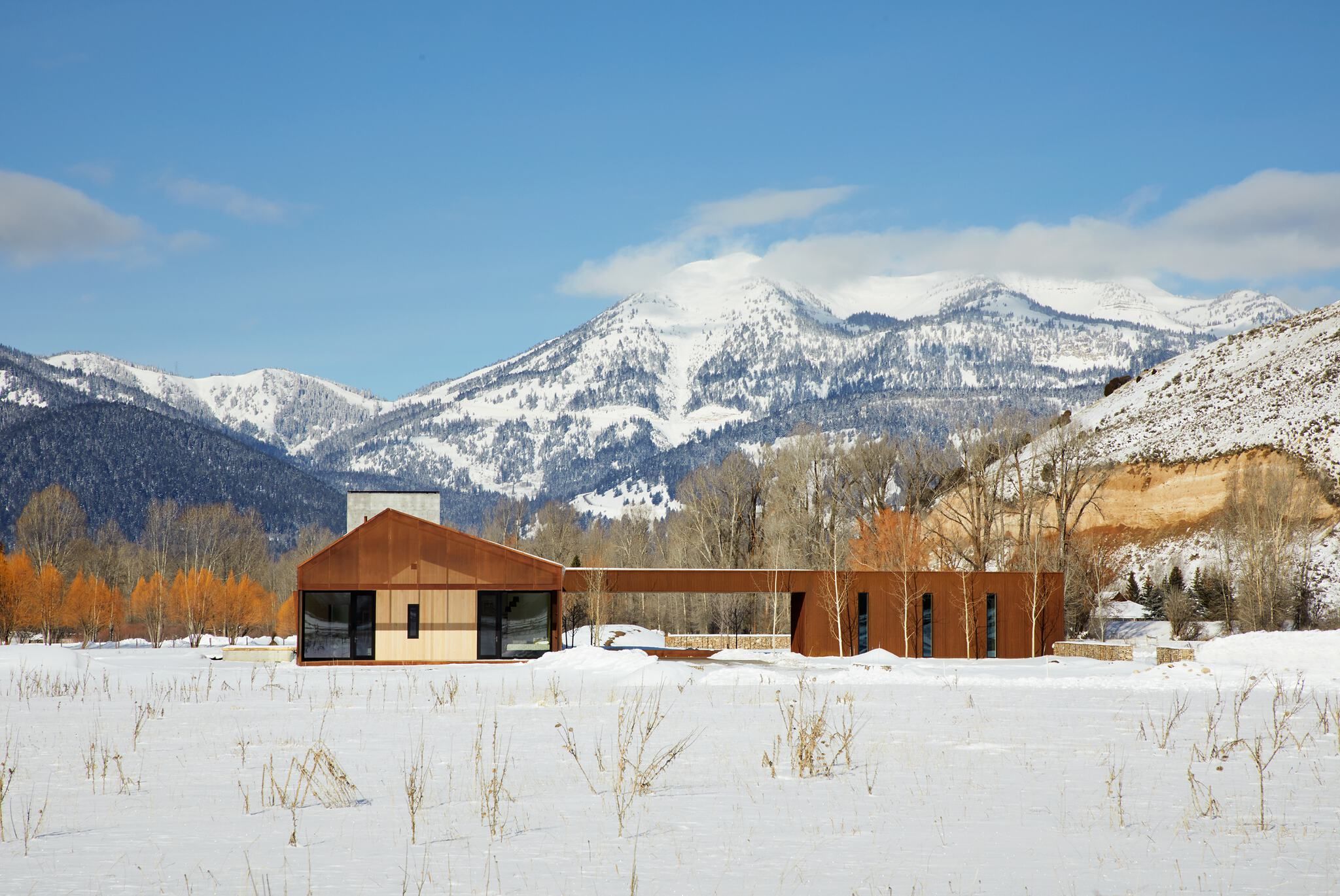 Contemporary Corten steel and wood house set in a snowy winter landscape with snow-capped mountains, evergreen forests, and a prominent tan cliff under a clear blue sky.