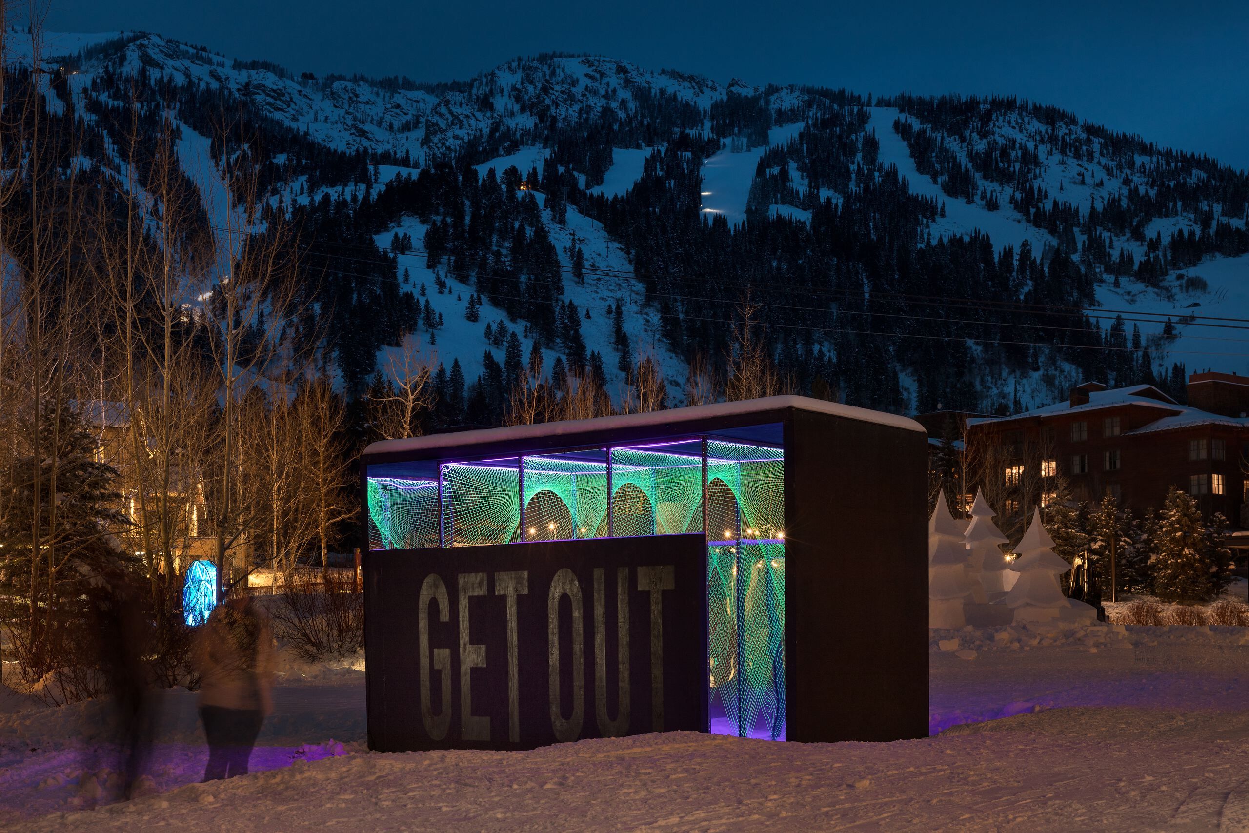 Nighttime winter scene featuring a GET OUT light art installation with glowing green and purple string patterns, set against snow-covered ski mountains and dark evergreen trees.