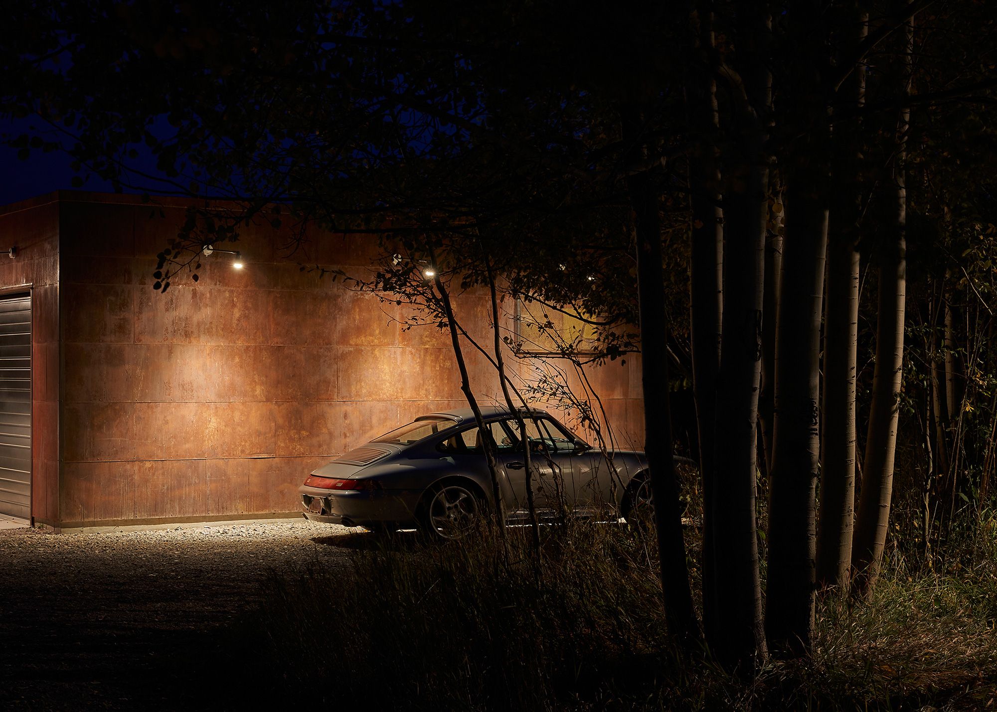Night view of a sports car parked beside an illuminated rusted steel building, framed by dark tree trunks.