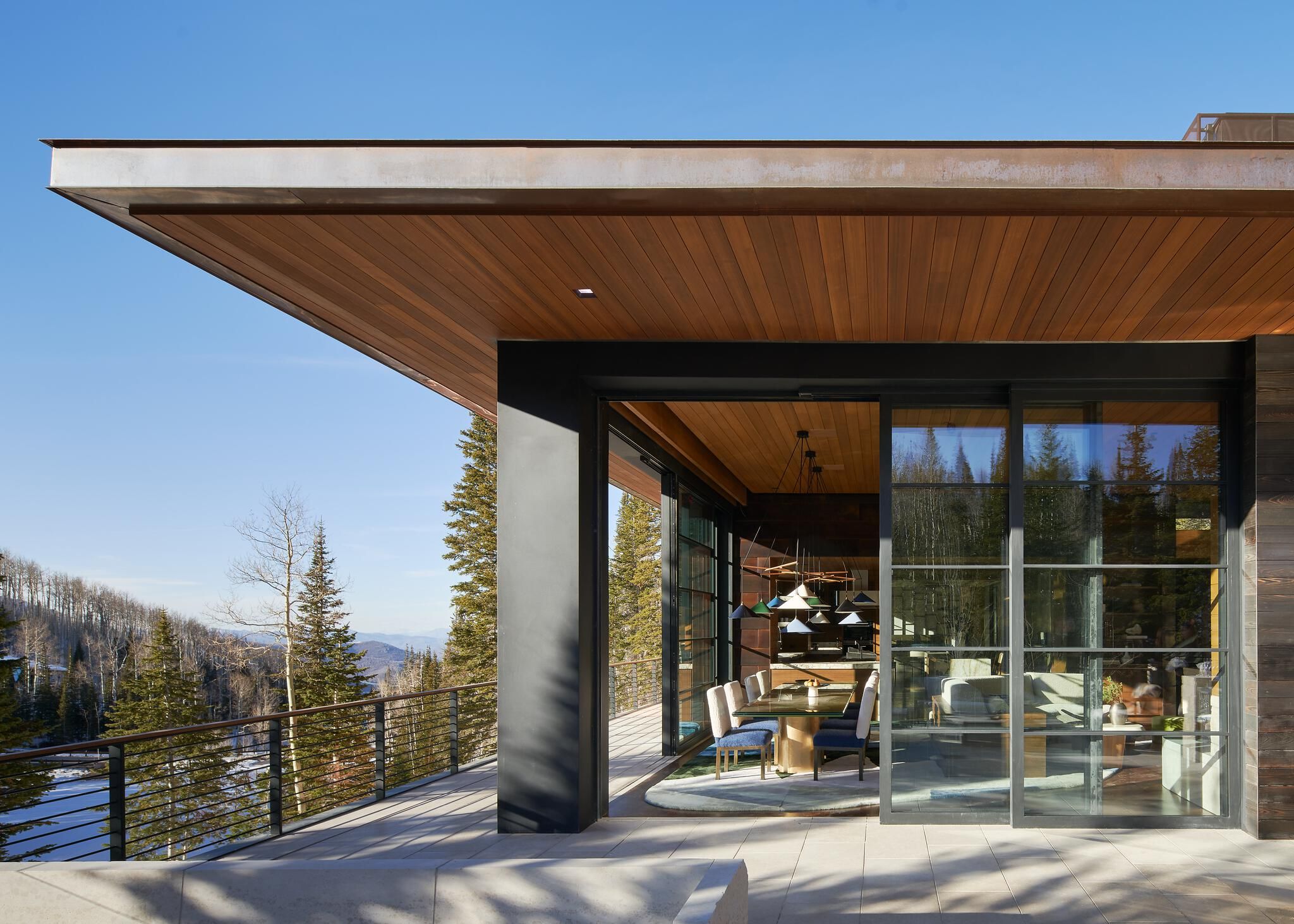 Modern mountain residence with an expansive deck and metal railing overlooking a snow-dusted forest and distant mountains. Large glass doors reveal a modern dining room inside, while a warm wood ceiling extends outdoors, complemented by dark structural elements under a clear blue sky.