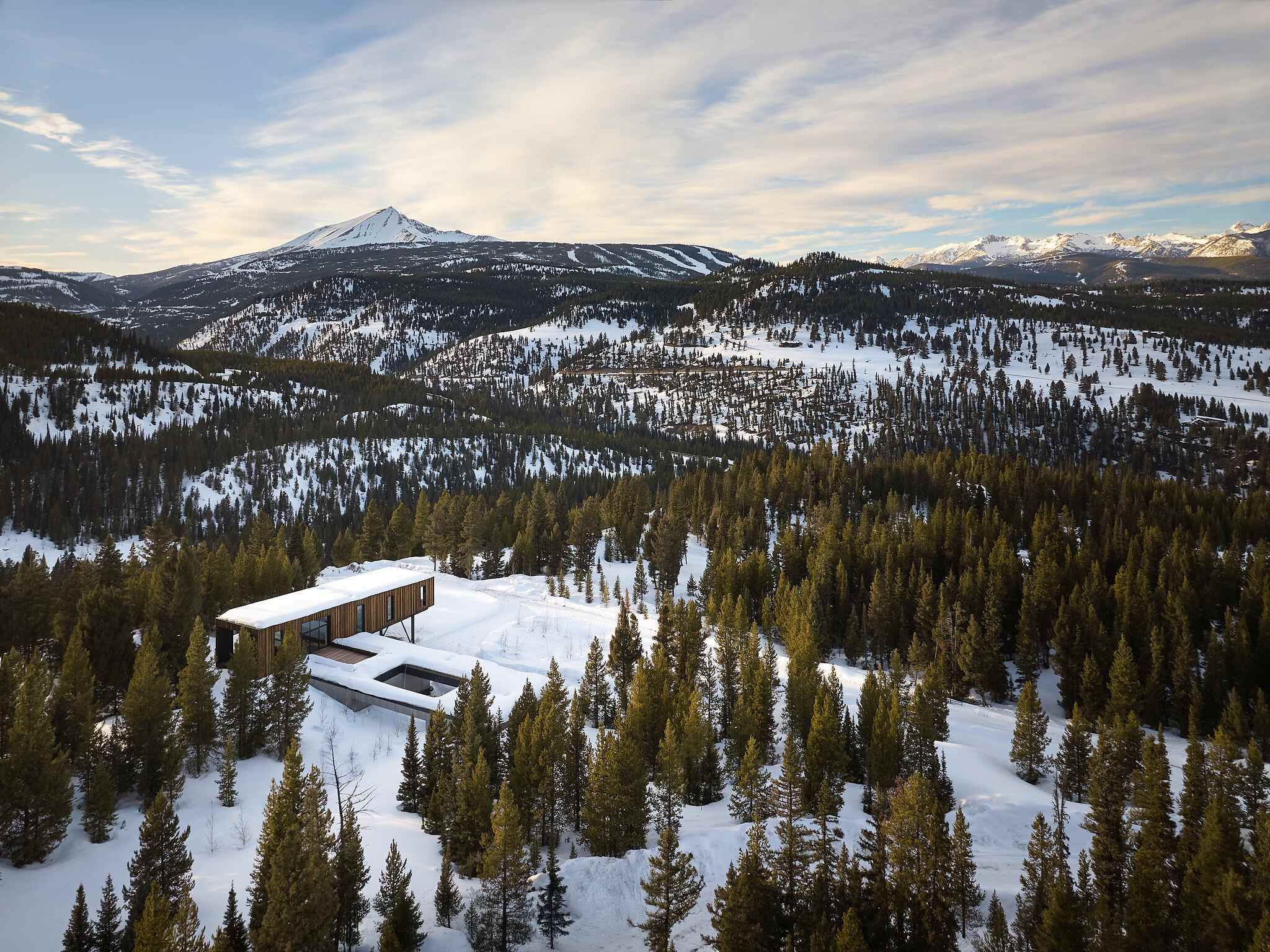 A striking modern architectural home, featuring warm wood siding and large glass windows, cantilevers over a snowy landscape with an integrated outdoor pool. It's nestled among dense evergreen trees, with majestic snow-capped mountains and distant ski slopes visible under a bright, partly cloudy sky.
