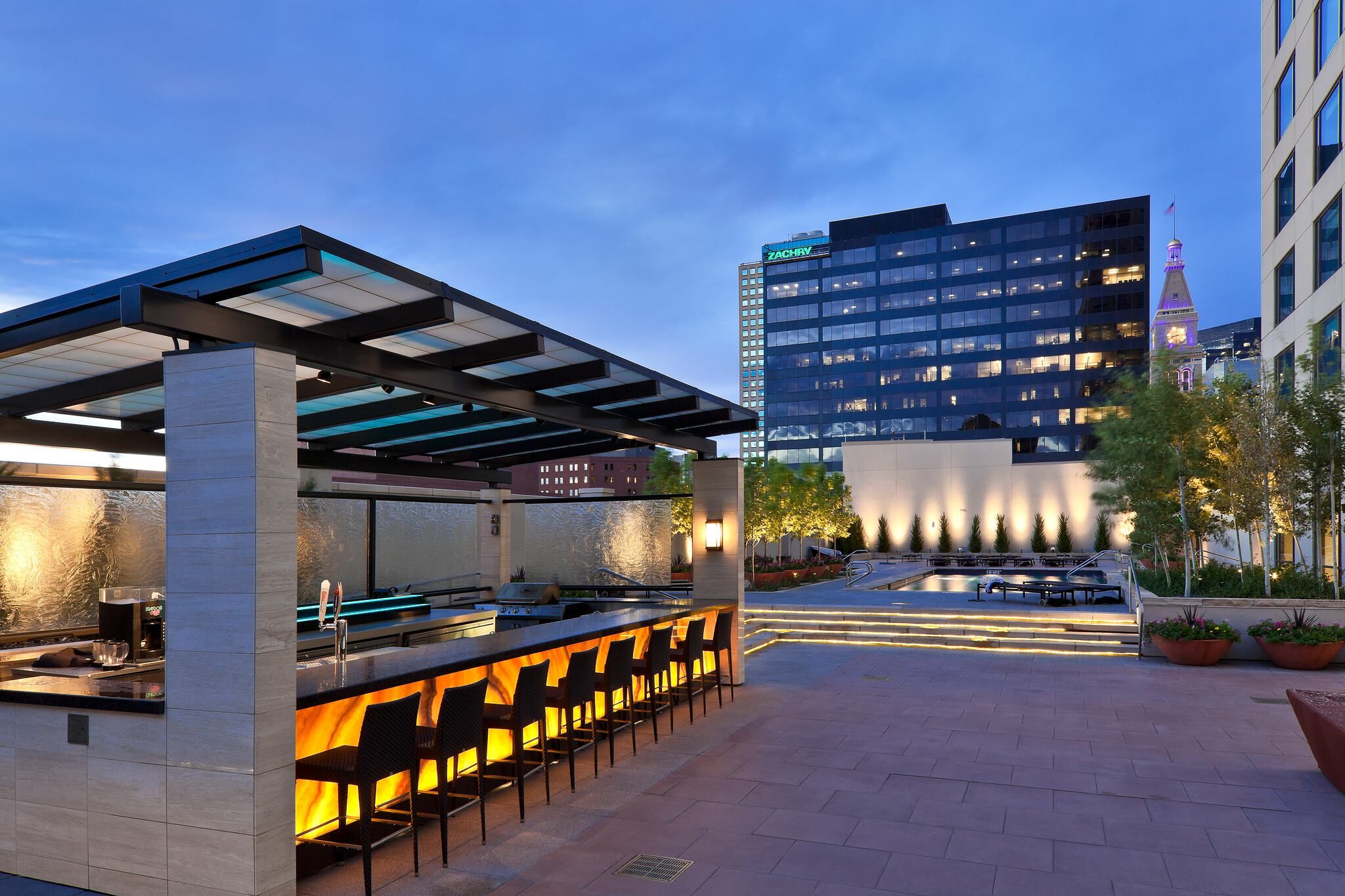 Modern rooftop bar and lounge at twilight, featuring an illuminated bar counter under a pergola, a pool deck with accent lighting, and a city skyline including a clock tower in the background.
