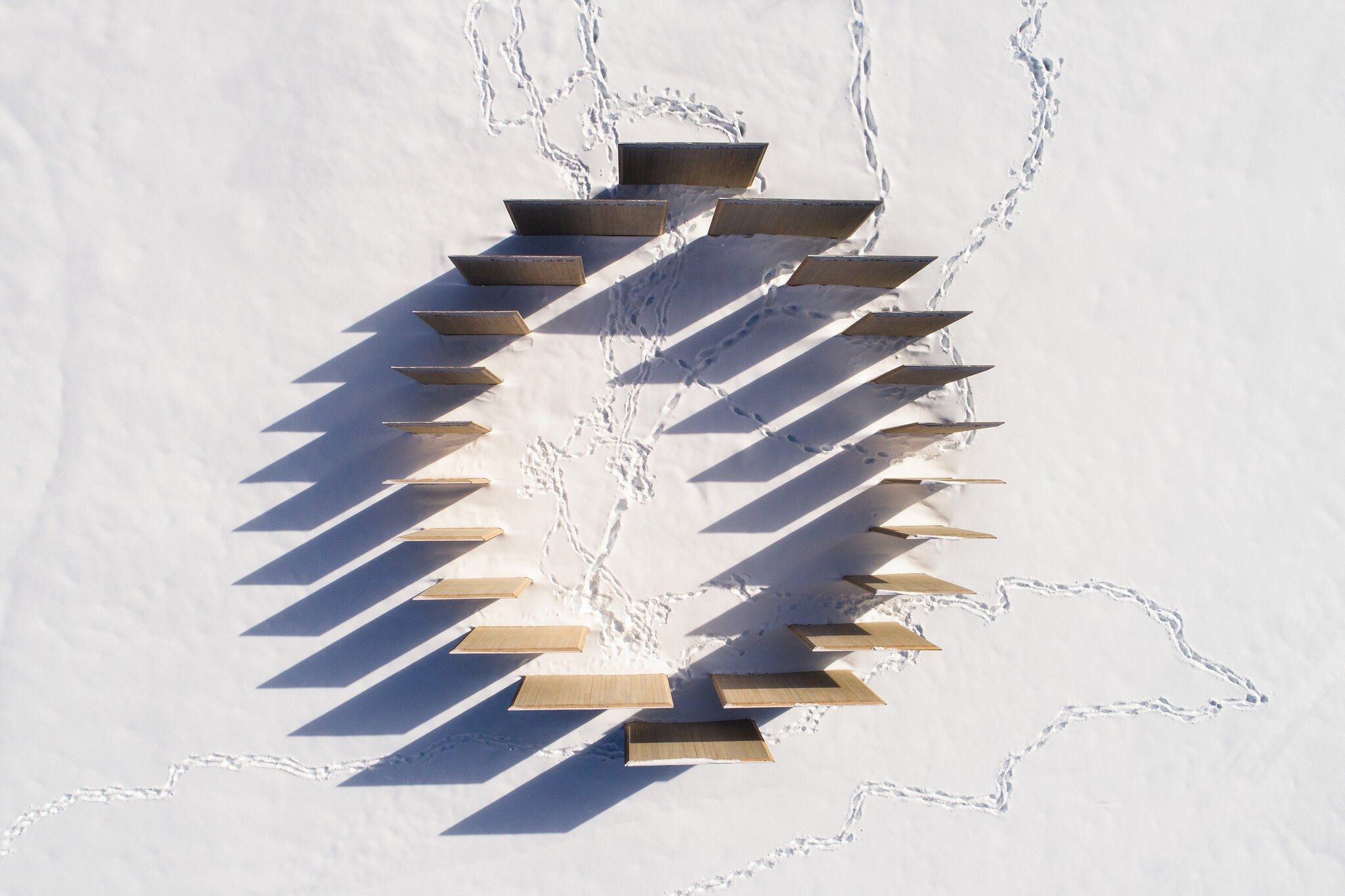 Aerial view of wooden platforms arranged in a radial pattern on fresh snow, casting long, sharp shadows, with numerous footprints winding between the structures.