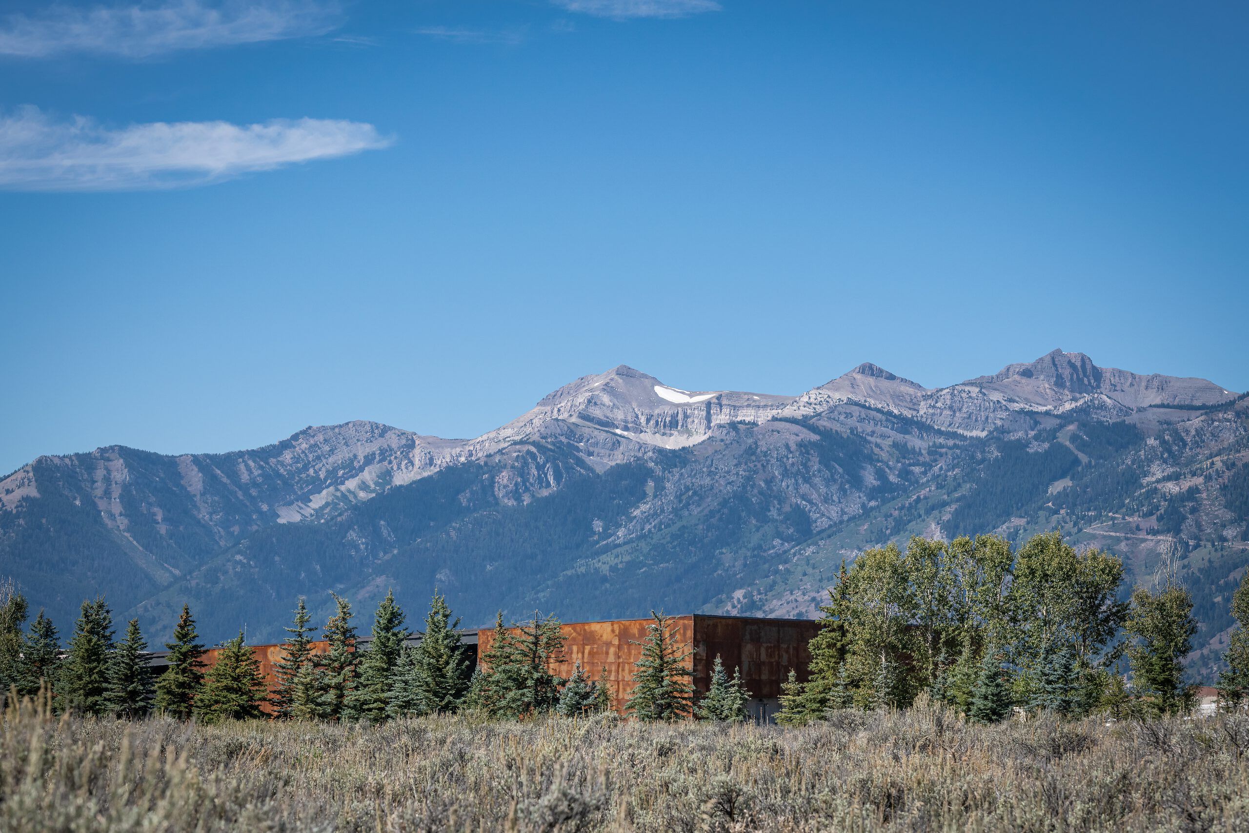 Dramatic mountain range with snow-dusted peaks and forested slopes under a clear blue sky. A modern, rectangular building with weathered steel or rust-colored walls is situated in the midground, partially concealed by evergreen trees and surrounded by dry, brushy vegetation.