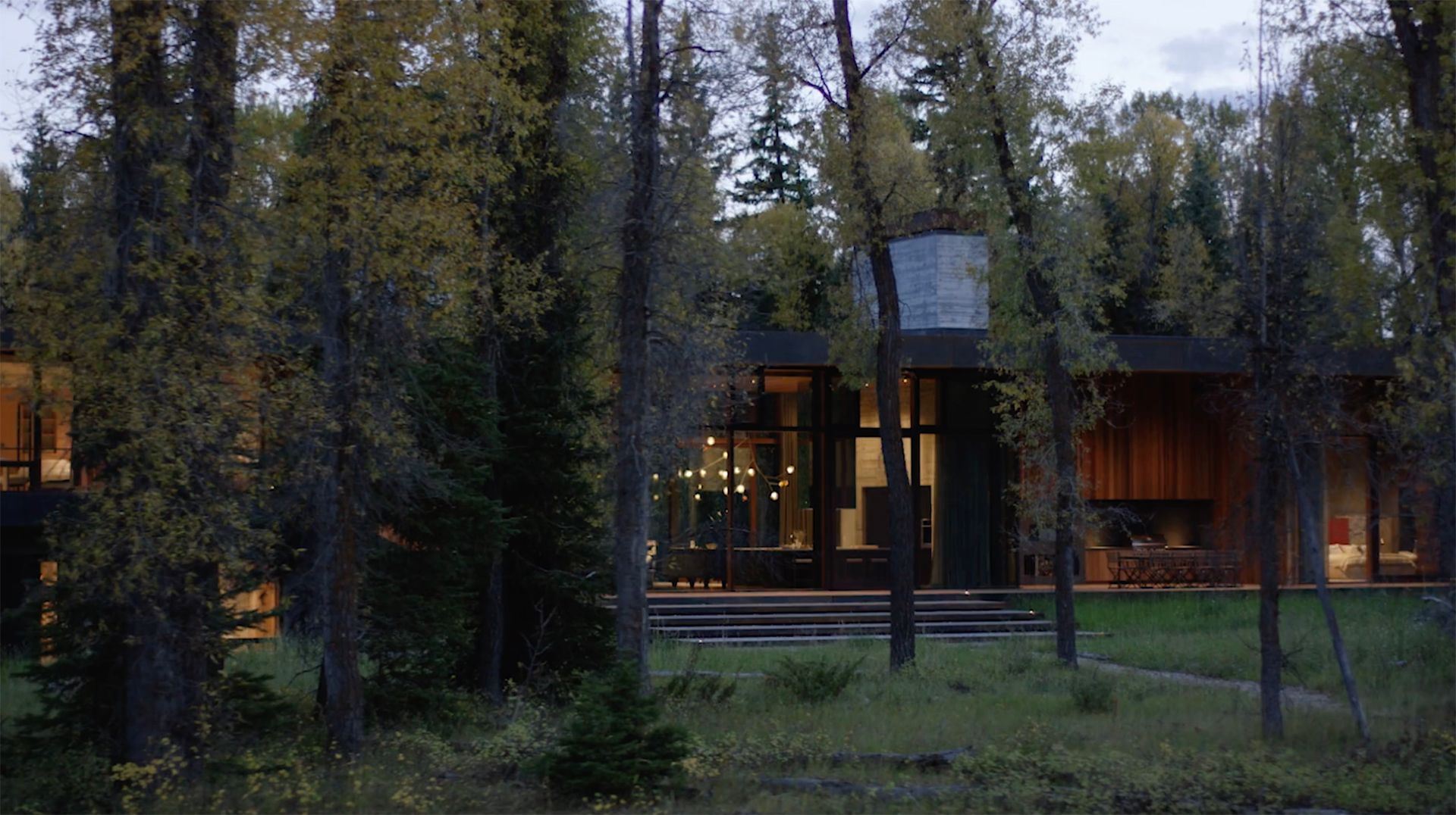 Modern architectural building at dusk, featuring vertical wooden siding, dark concrete, and horizontal wood panels, with warm light glowing from a tall window. Blurry tall grasses are in the foreground. A Vimeo video player overlay displays controls and the title CLB | Riverbend.