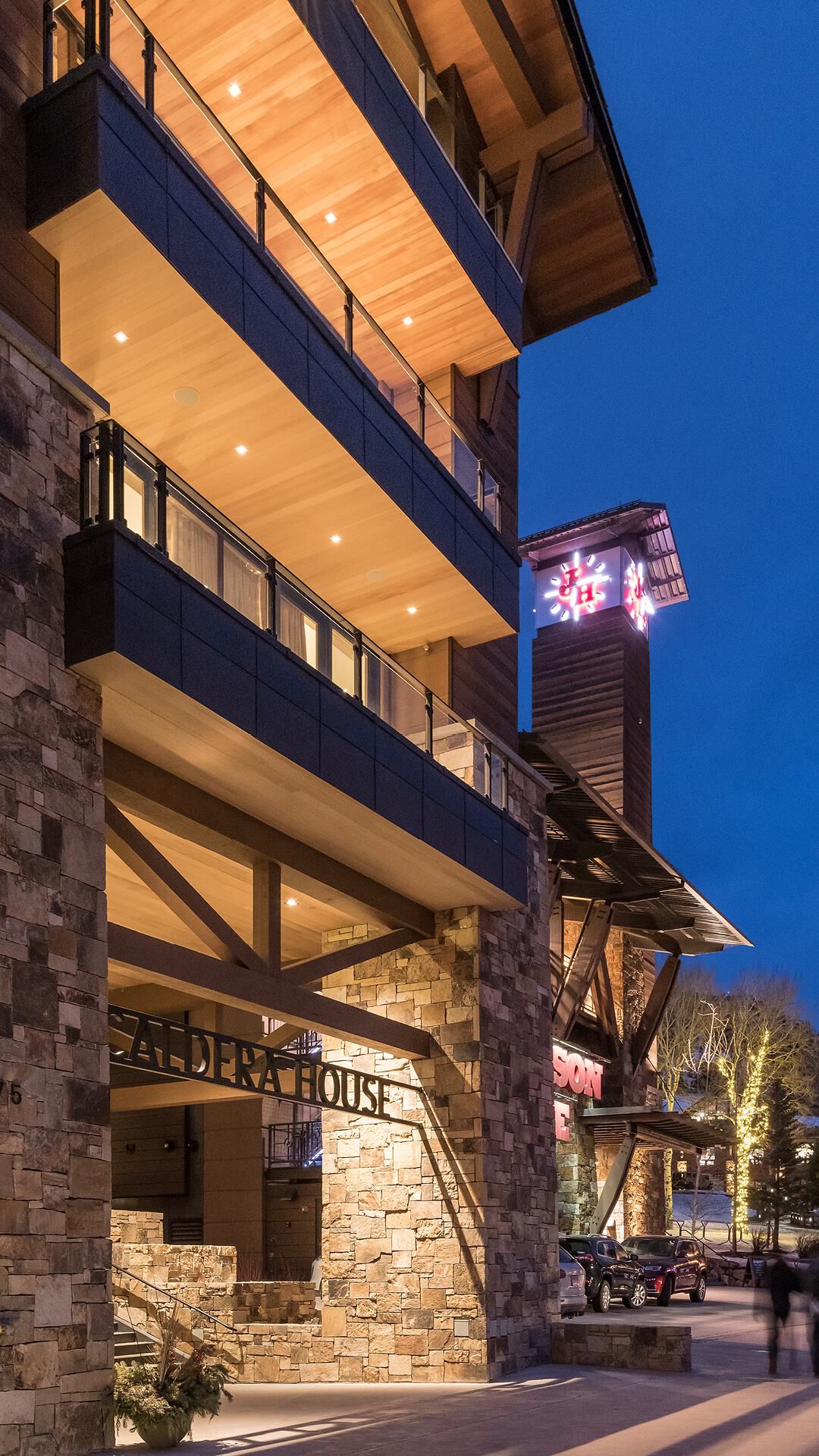 Modern Caldera House hotel at twilight, showcasing its stone and wood facade, illuminated balconies with glass railings, and prominent Caldera House entrance sign. A glowing clock tower with a red snowflake logo and parked cars are visible, suggesting a vibrant evening scene.