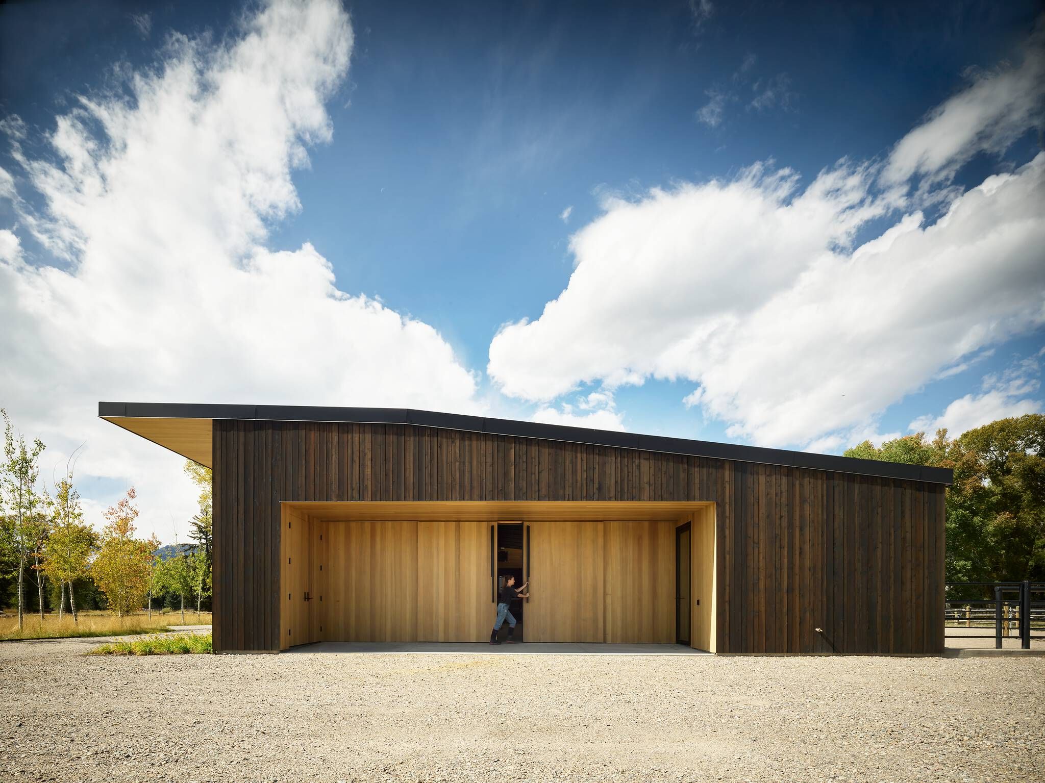 Modern stable with dark vertical wood siding and a black shed roof. A person opens one of the large, light wood sliding doors in a recessed entrance, facing a gravel foreground, autumnal trees, and a blue sky with white clouds.