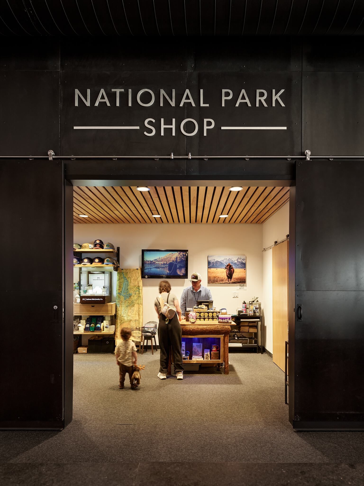 The NATIONAL PARK SHOP sign in white letters hangs above the open dark entrance. Inside, a brightly lit retail space features a woman and small child browsing, with a man working behind a rustic wooden counter. The shop interior includes a wooden slatted ceiling, displays of park-themed merchandise, and wall-mounted screens showing nature scenes. A toddler walks on the gray carpet, holding a toy deer.
