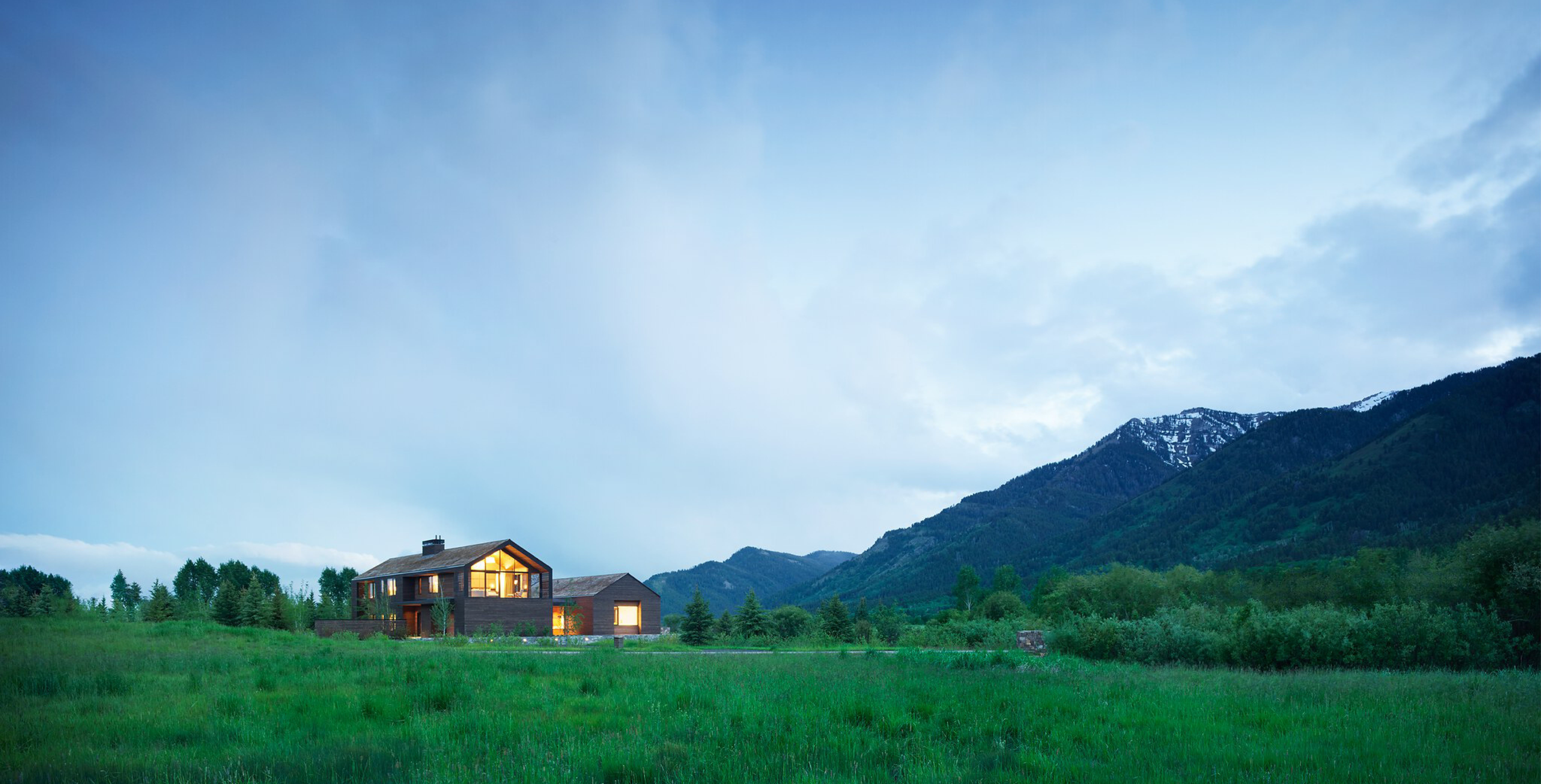 Modern dark wood house with warm lights, set in a lush green meadow, against a backdrop of forested mountains with snowy peaks under a twilight sky.