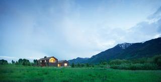 Modern dark wood house with warm lights, set in a lush green meadow, against a backdrop of forested mountains with snowy peaks under a twilight sky.
