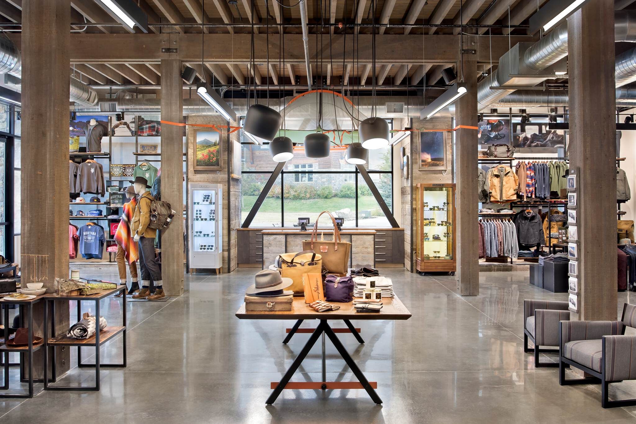 Modern rustic retail store interior with an open layout, showcasing lifestyle clothing and accessories on mannequins and display tables. The space features exposed wood beams, concrete floors, industrial lighting, and large windows with an outdoor view.