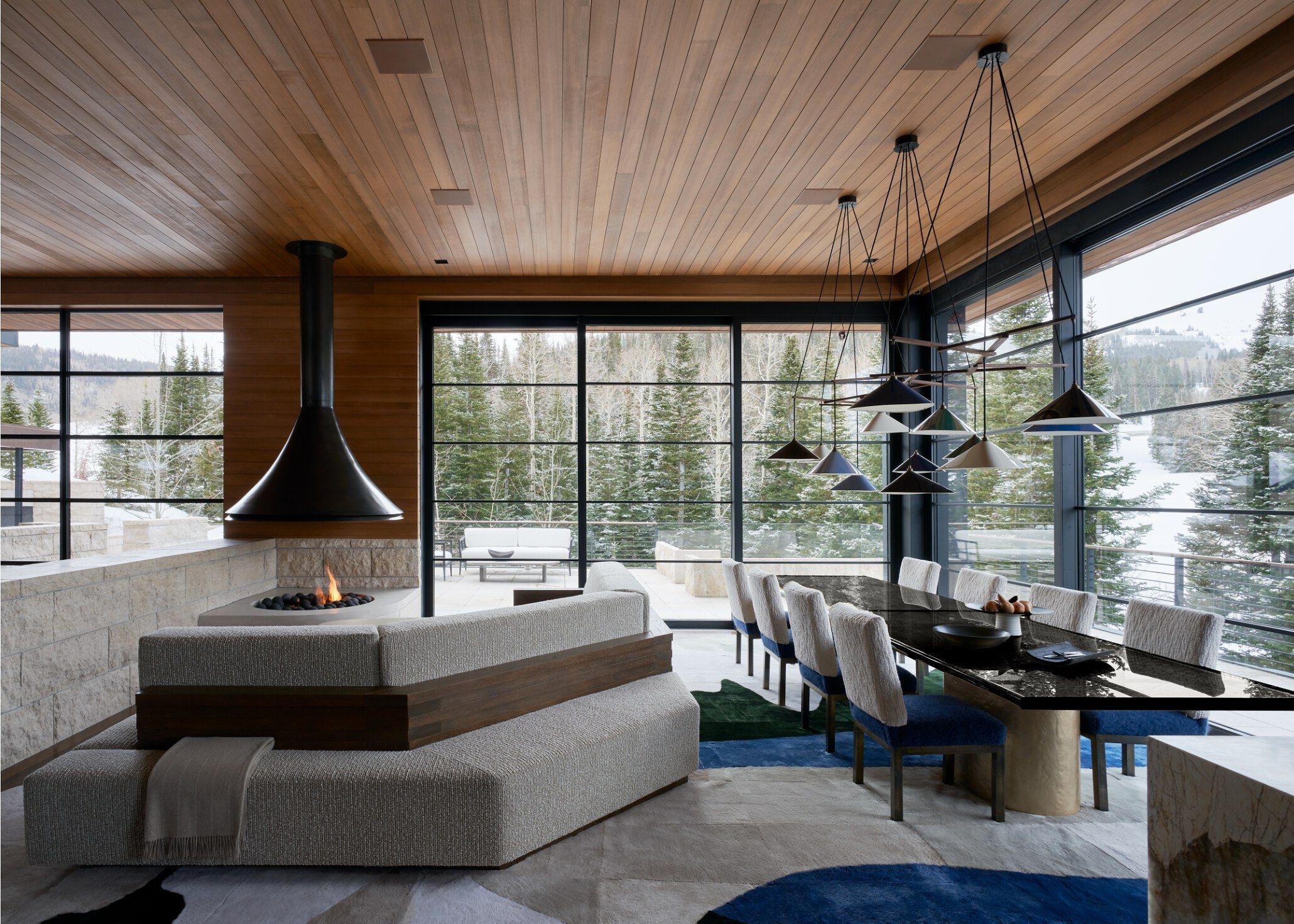 Contemporary open-plan dining and living room in a luxury mountain home, featuring a long black dining table with textured chairs and modern pendant lights. A grey sectional sofa faces a built-in fireplace with a black hood. Large windows showcase a stunning snowy forest and mountain landscape, complemented by a wood-paneled ceiling and stone accent wall.