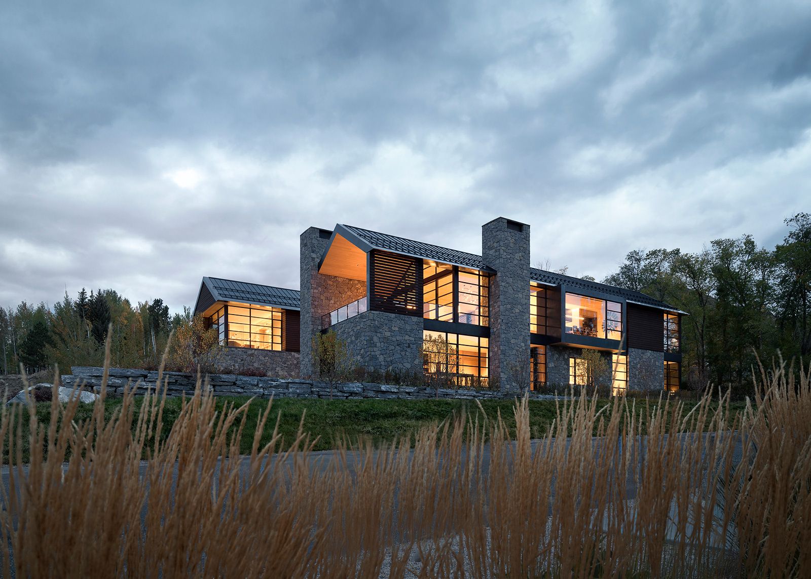 Evening view of a multi-story modern stone and glass mountain villa framed by tall ornamental grass in the foreground.