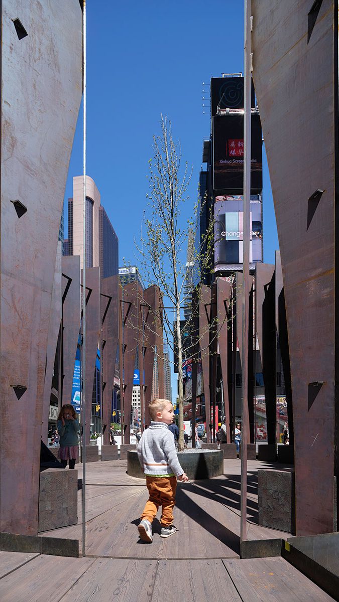 A young boy walks on a wooden path amidst an urban art installation featuring tall, weathered metal panels. Mirrored panels reflect the boy, surrounding city buildings, and bright digital billboards under a clear blue sky.
