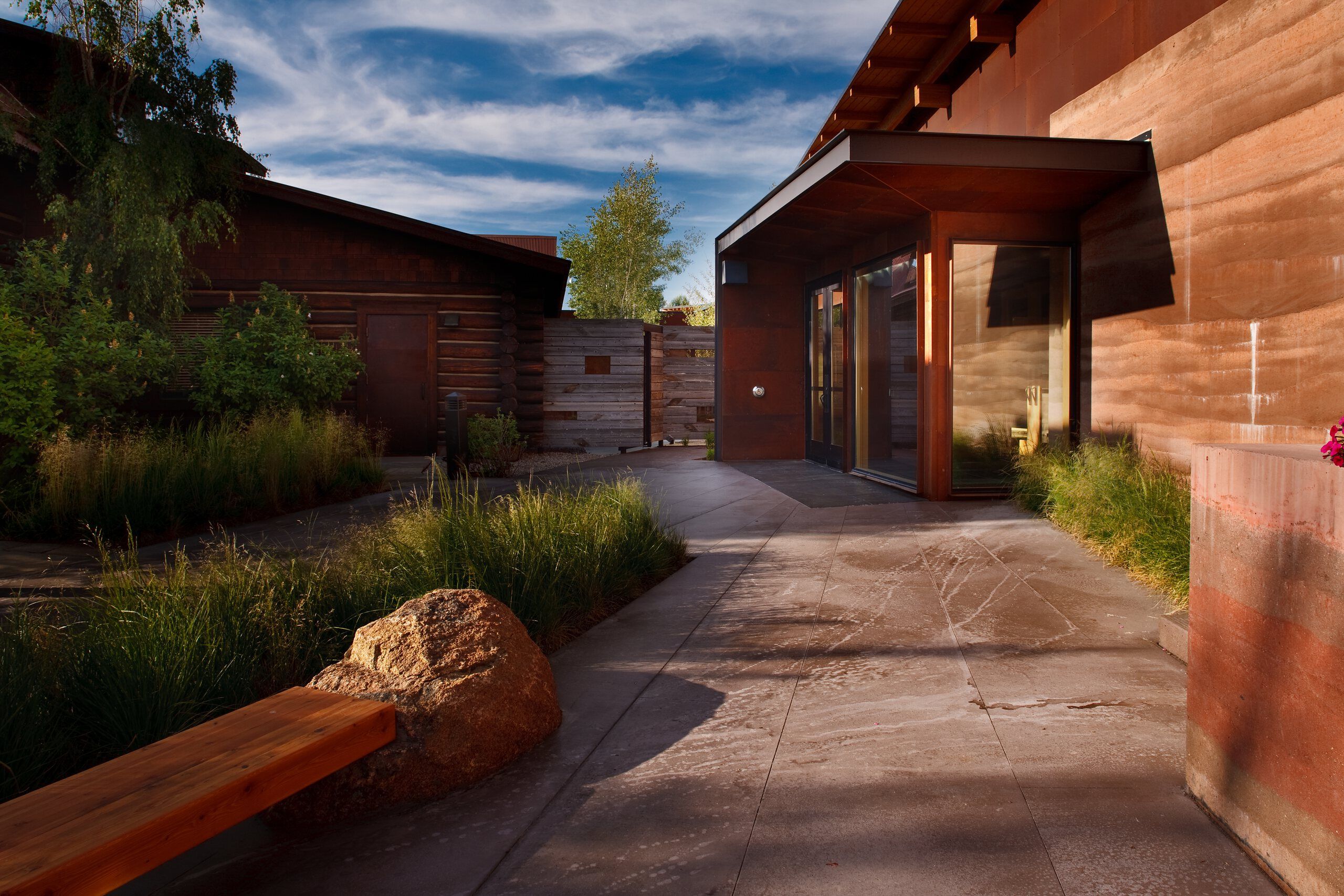 A sunny exterior view showcasing a rustic log cabin next to a contemporary building with rusted metal and layered rammed earth walls. A stone pathway leads between them, bordered by lush grasses, a large boulder, and a wooden bench, all casting long shadows under a blue, cloudy sky.