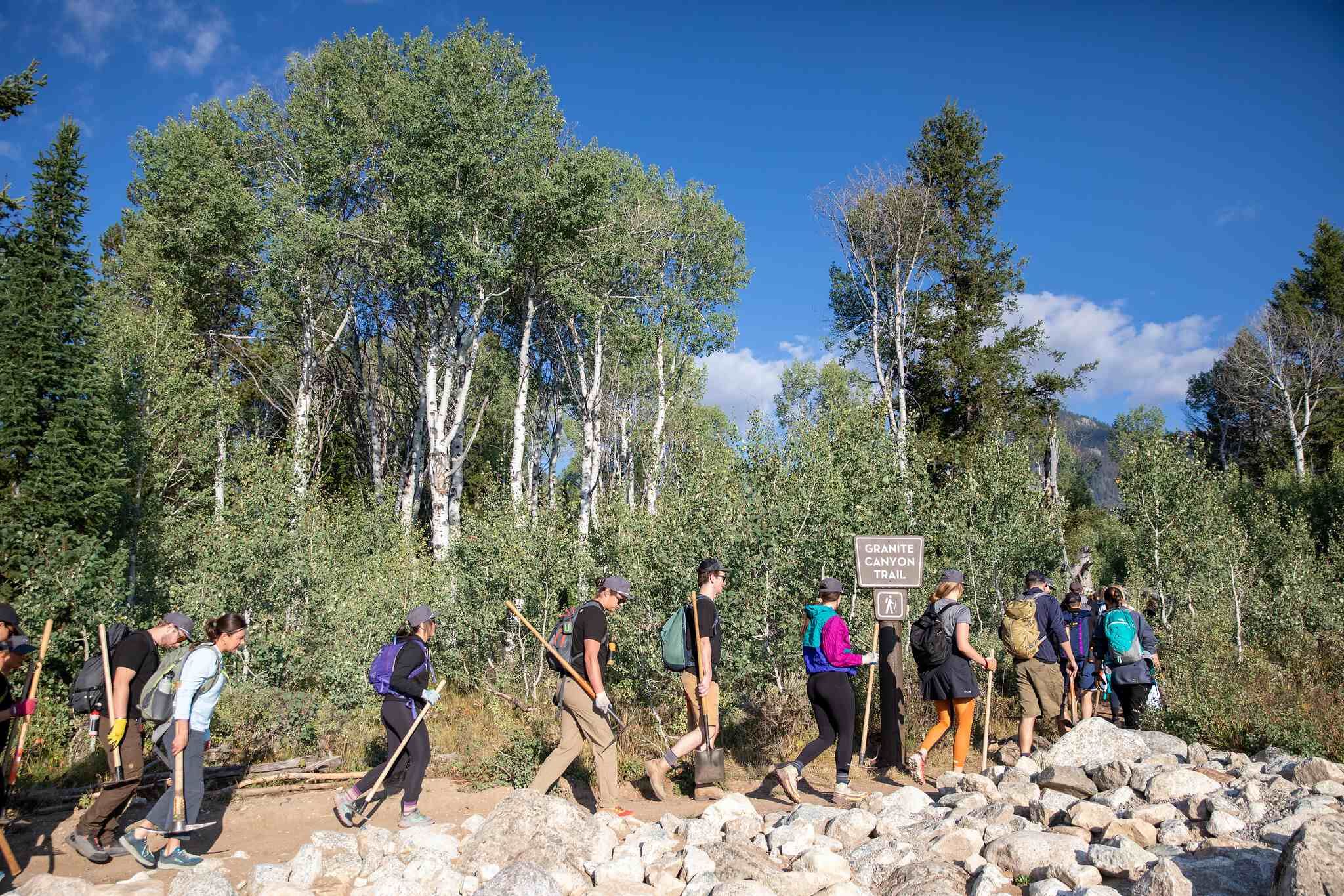 Group of people carrying trail maintenance tools and backpacks walking on a rocky path labeled Granite Canyon Trail through a forest of aspen and evergreen trees under a clear blue sky.