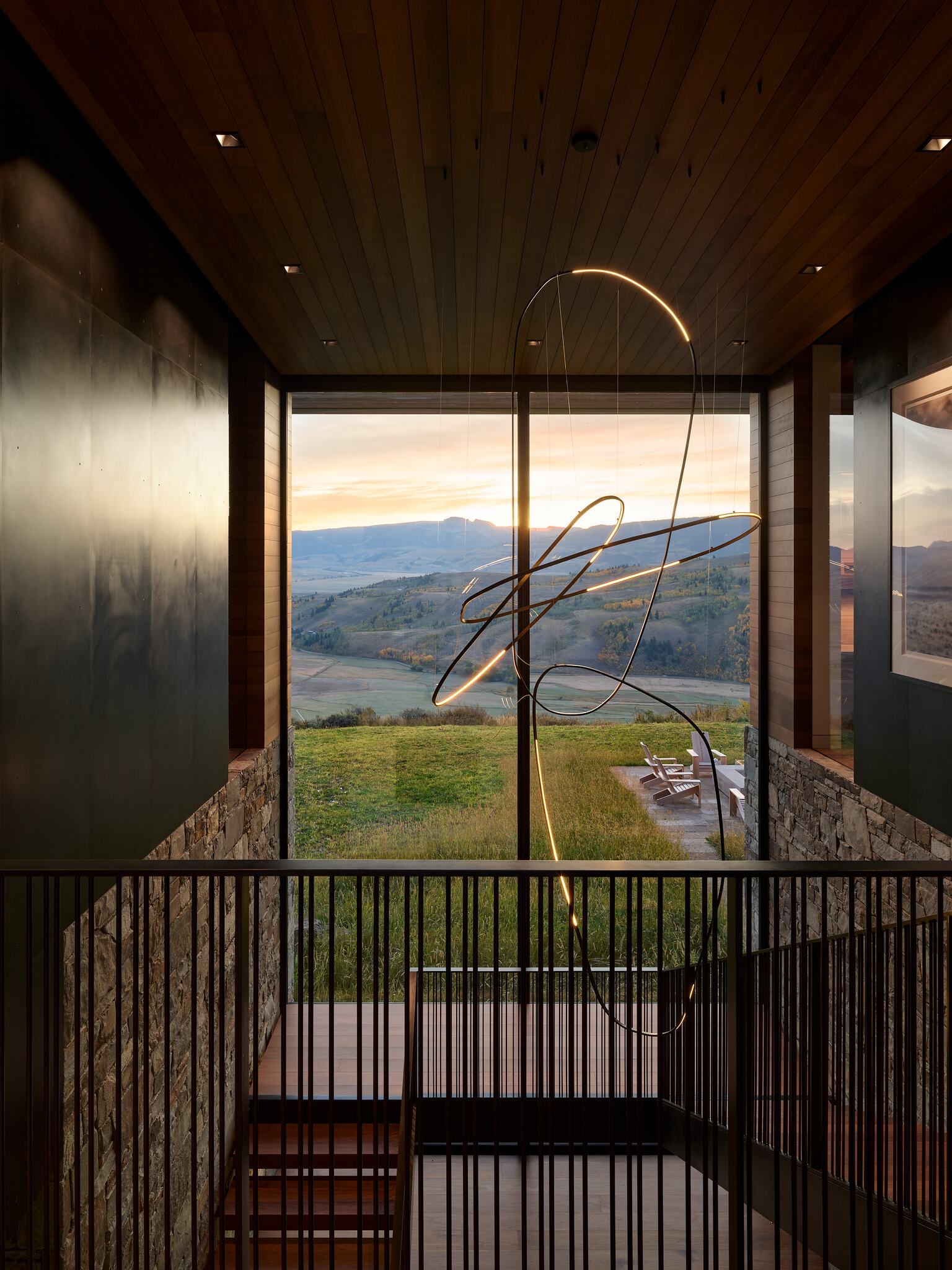 A contemporary multi-level interior features a large window with a panoramic view of a sunset over a rolling valley and mountains. A striking, abstract light sculpture with glowing curvilinear elements hangs prominently, reflecting the golden hour light. The interior showcases dark wood paneling on the ceiling, stone and metal-clad walls, and a minimalist metal railing leading down a staircase.