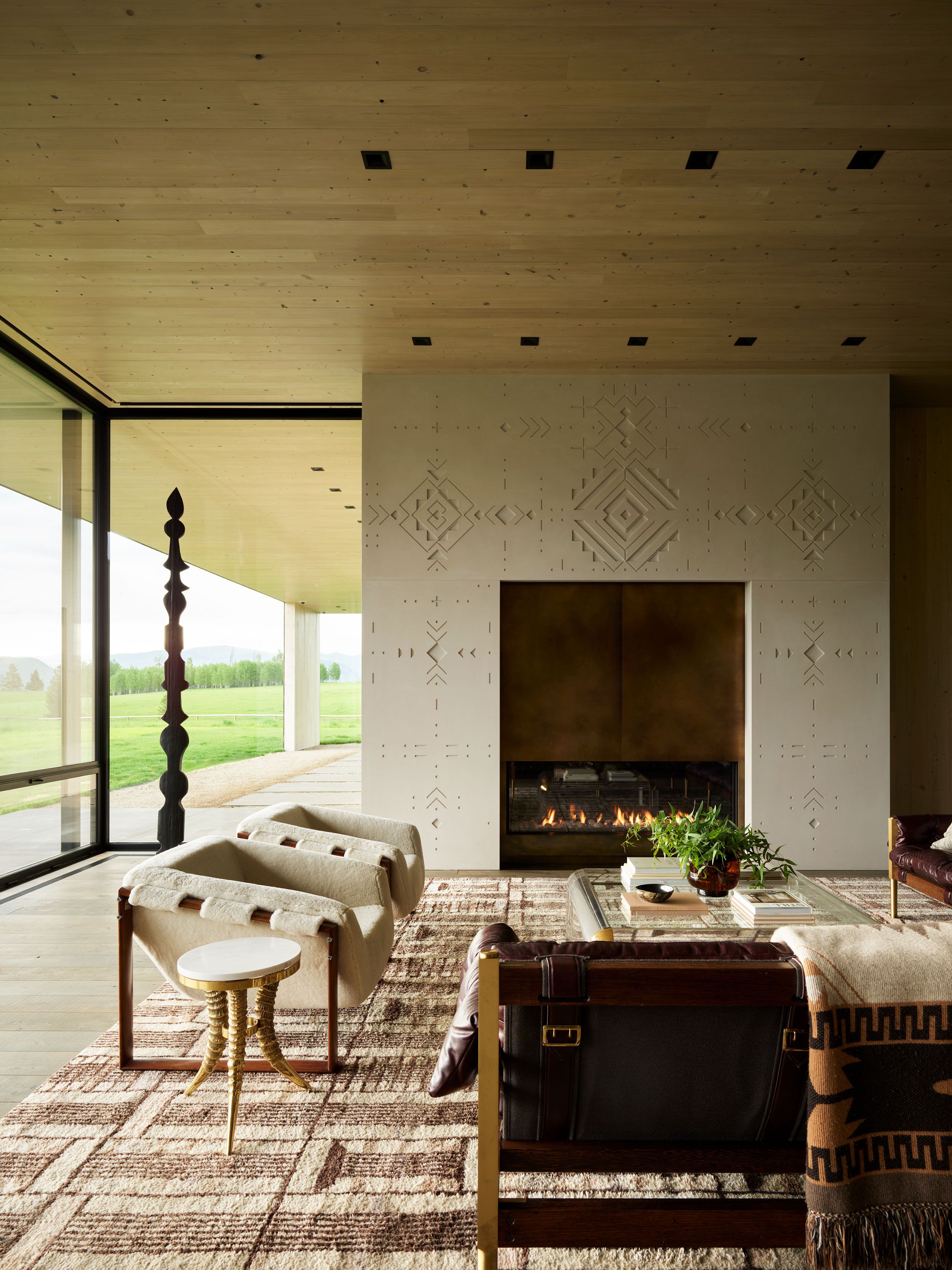 Sophisticated living room with a fireplace featuring intricate geometric wall patterns, a large glass coffee table, and comfortable seating. Expansive floor-to-ceiling windows overlook a vibrant green landscape. The room has light wood plank ceilings and a textured Southwestern-style rug.