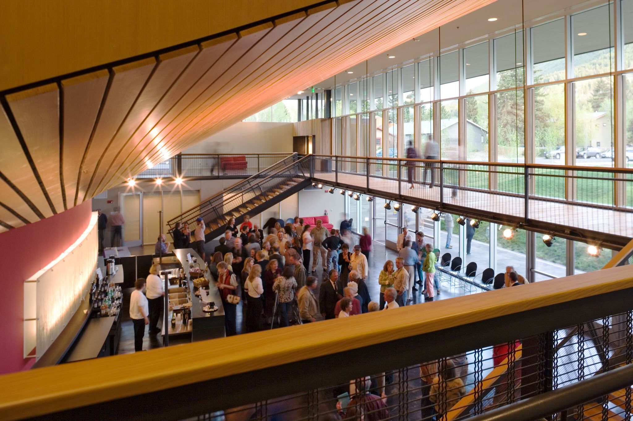 A modern, multi-level lobby filled with people mingling at an event, featuring high ceilings with warm wooden panels, expansive windows overlooking a forested outdoor area, a prominent staircase, an elevated walkway, and a bar on the lower level.