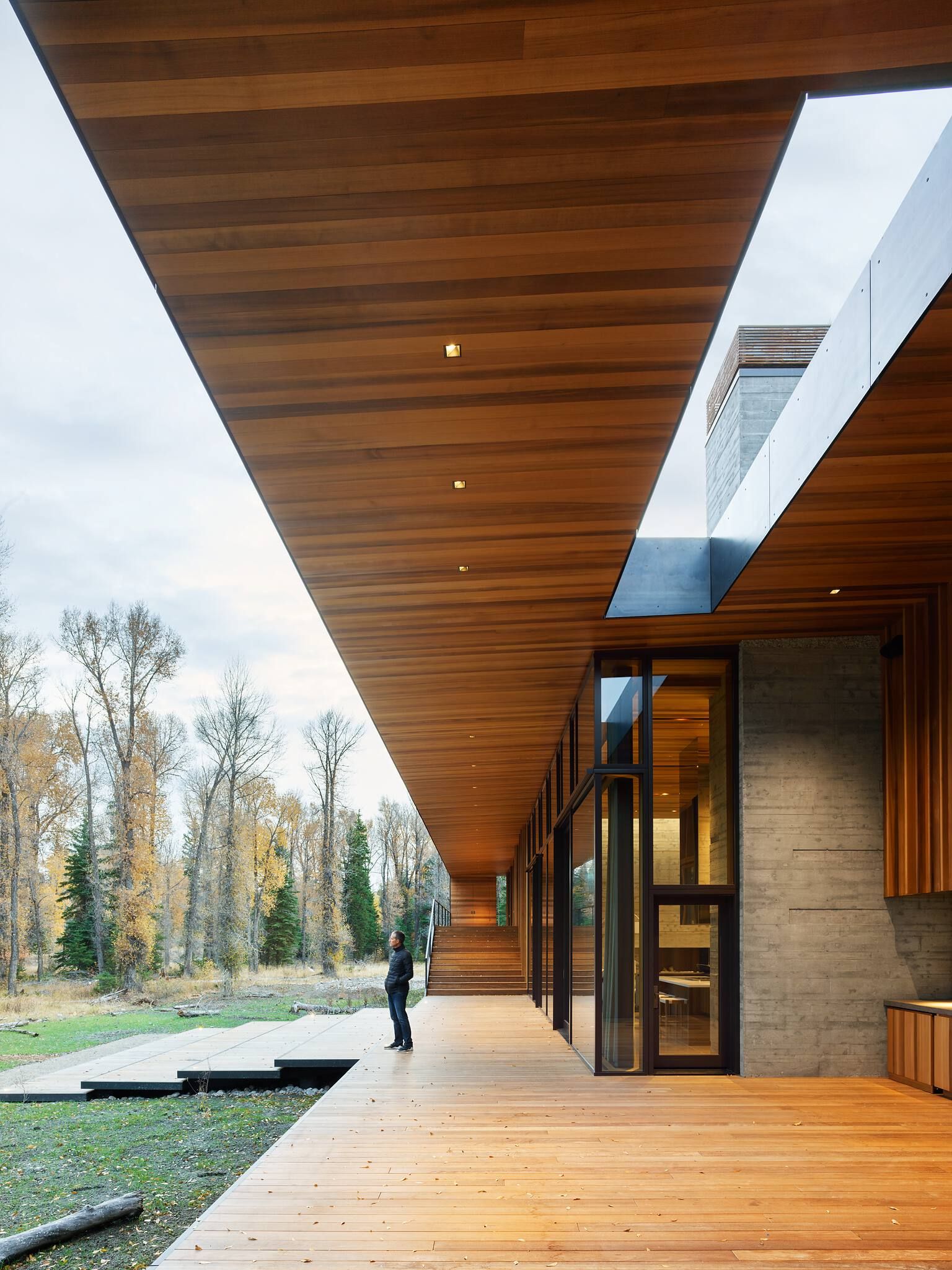 Modern luxury home with a long wooden deck and matching slatted ceiling, featuring floor-to-ceiling glass walls. A man stands on the deck looking out at the surrounding autumn trees and natural landscape, with concrete walls visible.