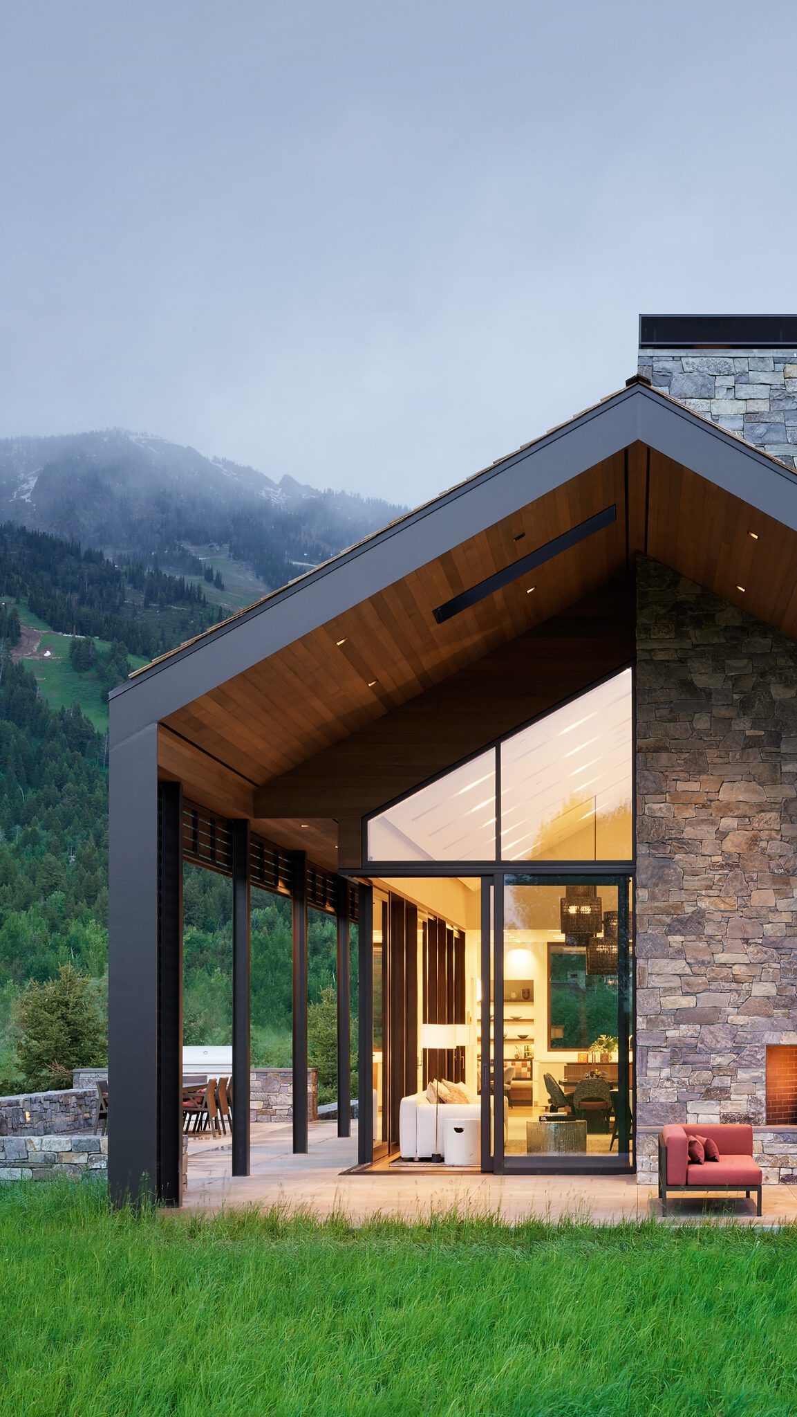 Modern mountain home with a stone exterior, expansive glass windows revealing an illuminated interior, and a covered patio with outdoor seating, set against a backdrop of misty, forested mountains at dusk.