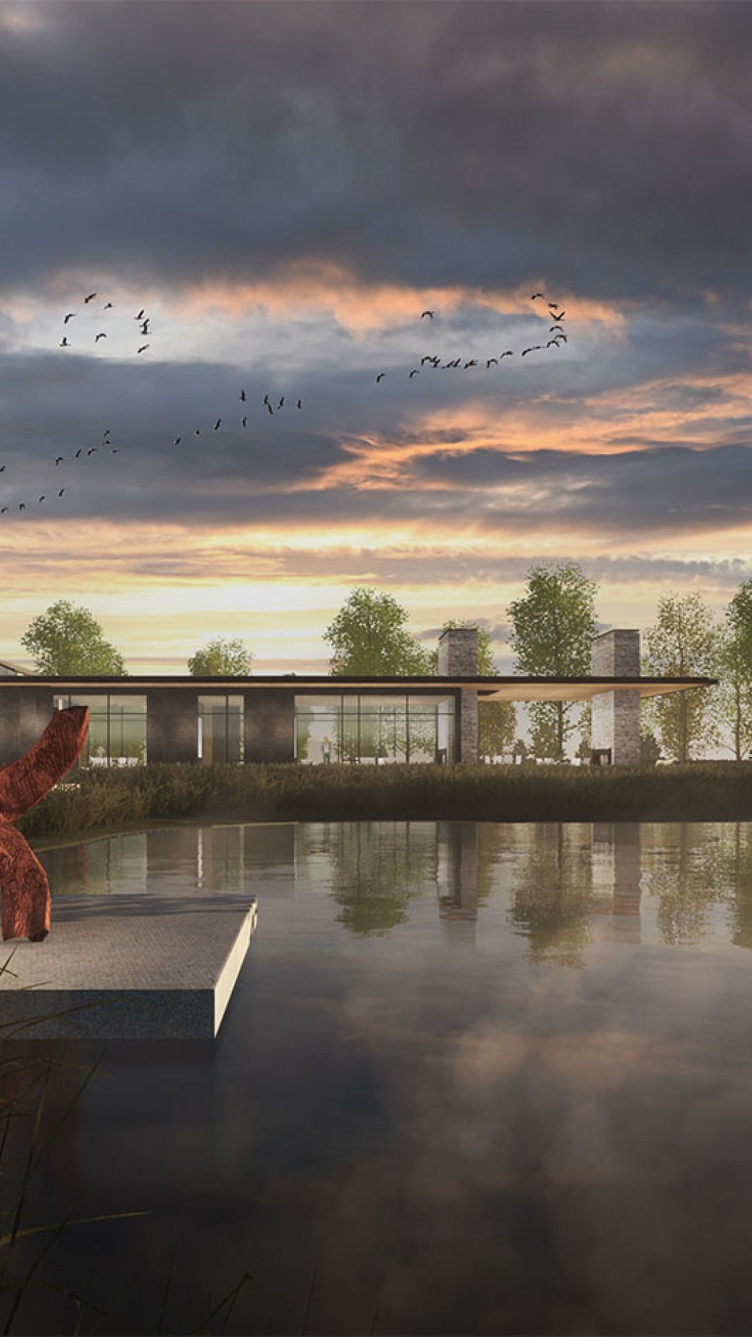 Modern architectural building with large glass windows and a dark facade by a tranquil lake. The dramatic sunset sky, featuring warm orange and pink clouds and flocks of flying birds, is reflected on the water's surface. An abstract reddish-brown sculpture rests on a concrete platform extending into the lake in the foreground.