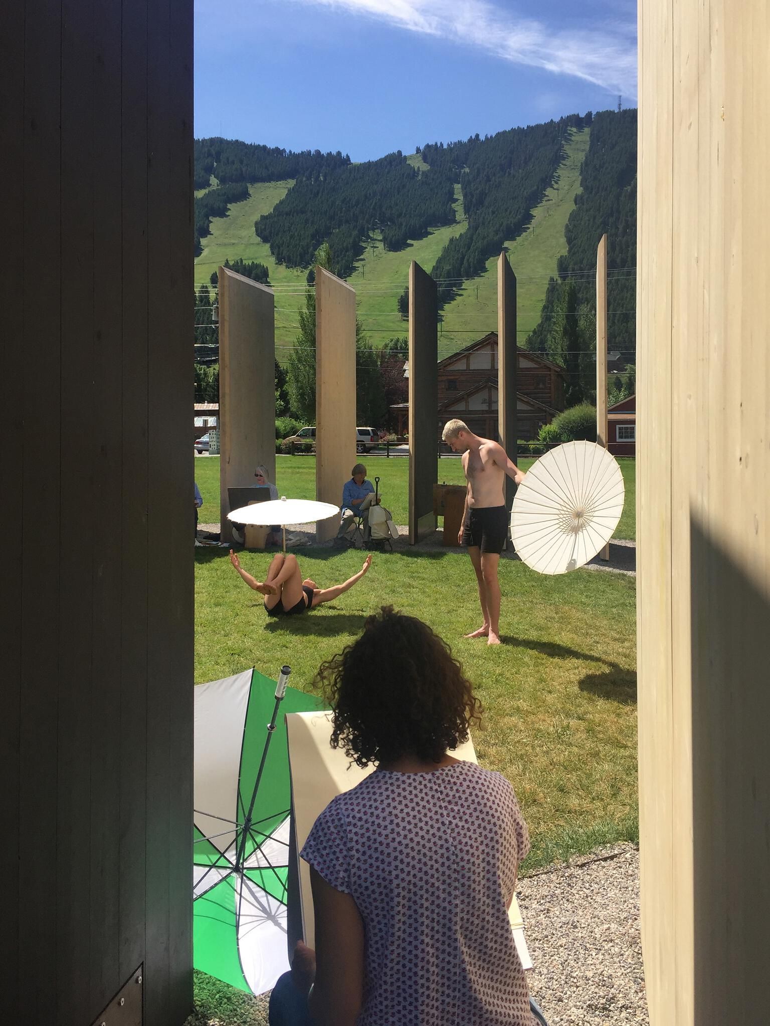 An outdoor figure drawing session on a sunny day with a woman posing on her back in the grass, a shirtless man holding a large white parasol, and observers, set against a lush green mountain slope.