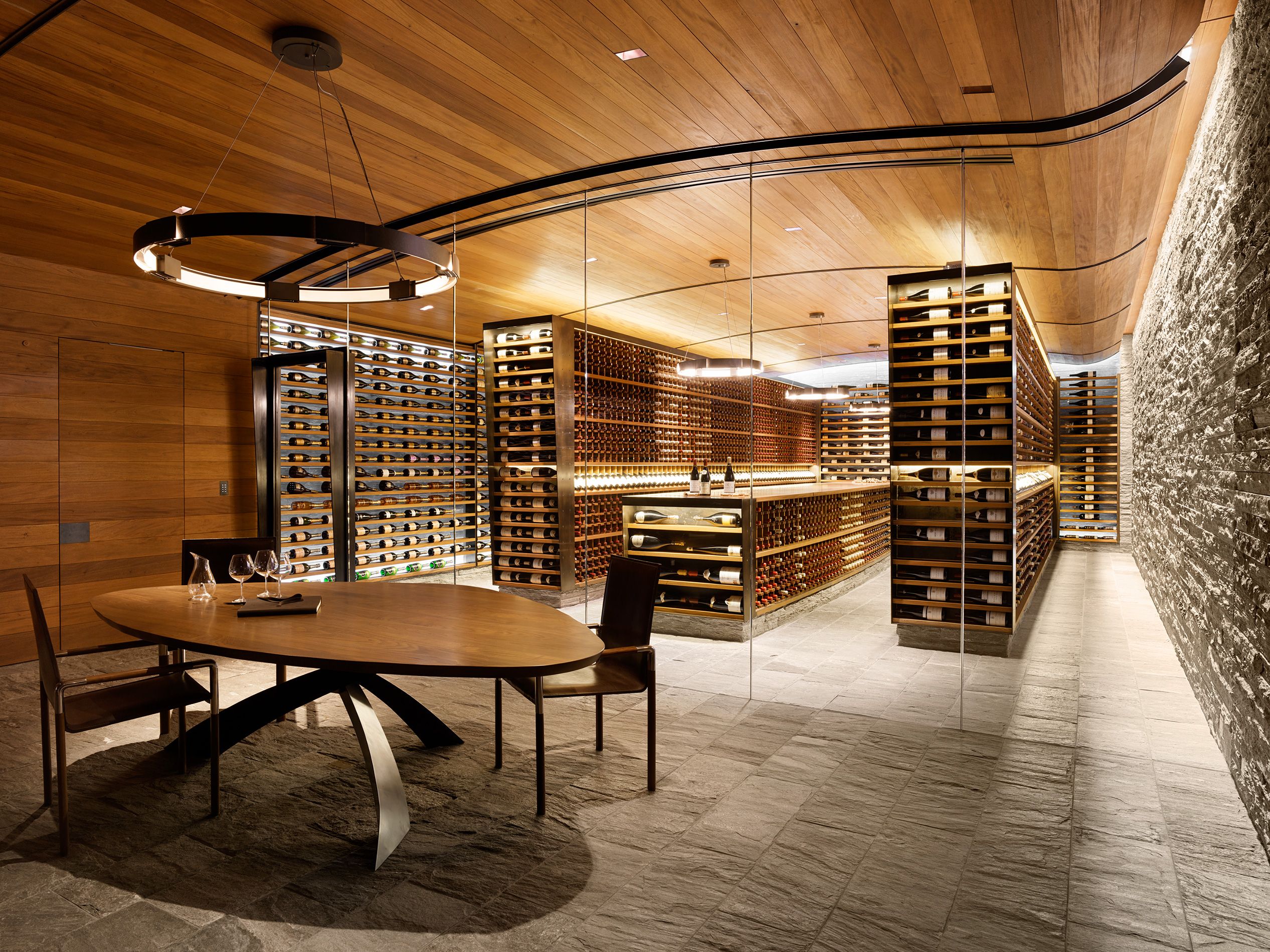Luxurious glass-enclosed wine cellar with custom lit racks, paired with an oval dining table set for tasting.