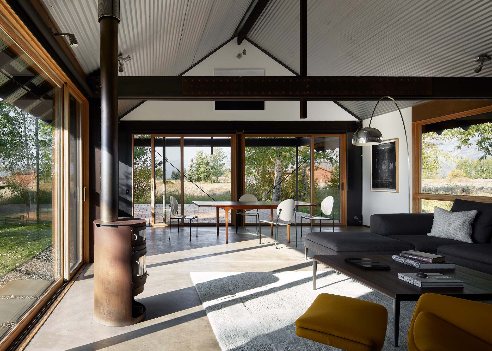 Modern open-plan living and dining room with a corrugated metal ceiling, exposed beams, and polished concrete floors. A rustic wood-burning stove stands by large glass doors offering views of a natural landscape. The living area features a dark sectional sofa, wood coffee table, and mustard yellow armchairs.