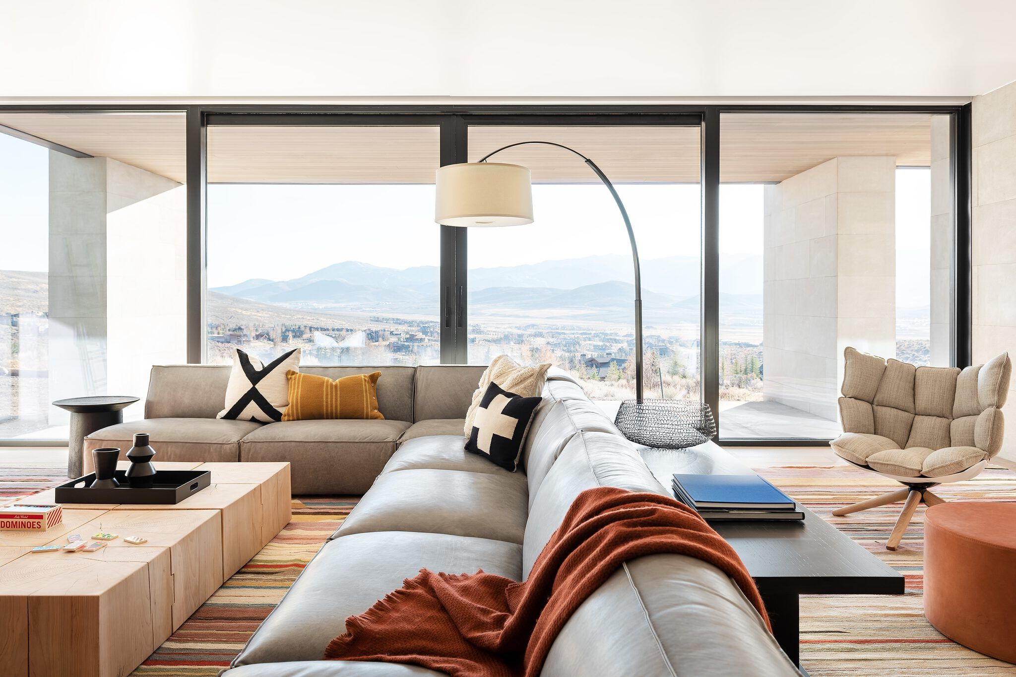 Spacious modern living room with a large light grey sectional sofa, wood block coffee tables, and a colorful rug, offering panoramic mountain and valley views through floor-to-ceiling windows.