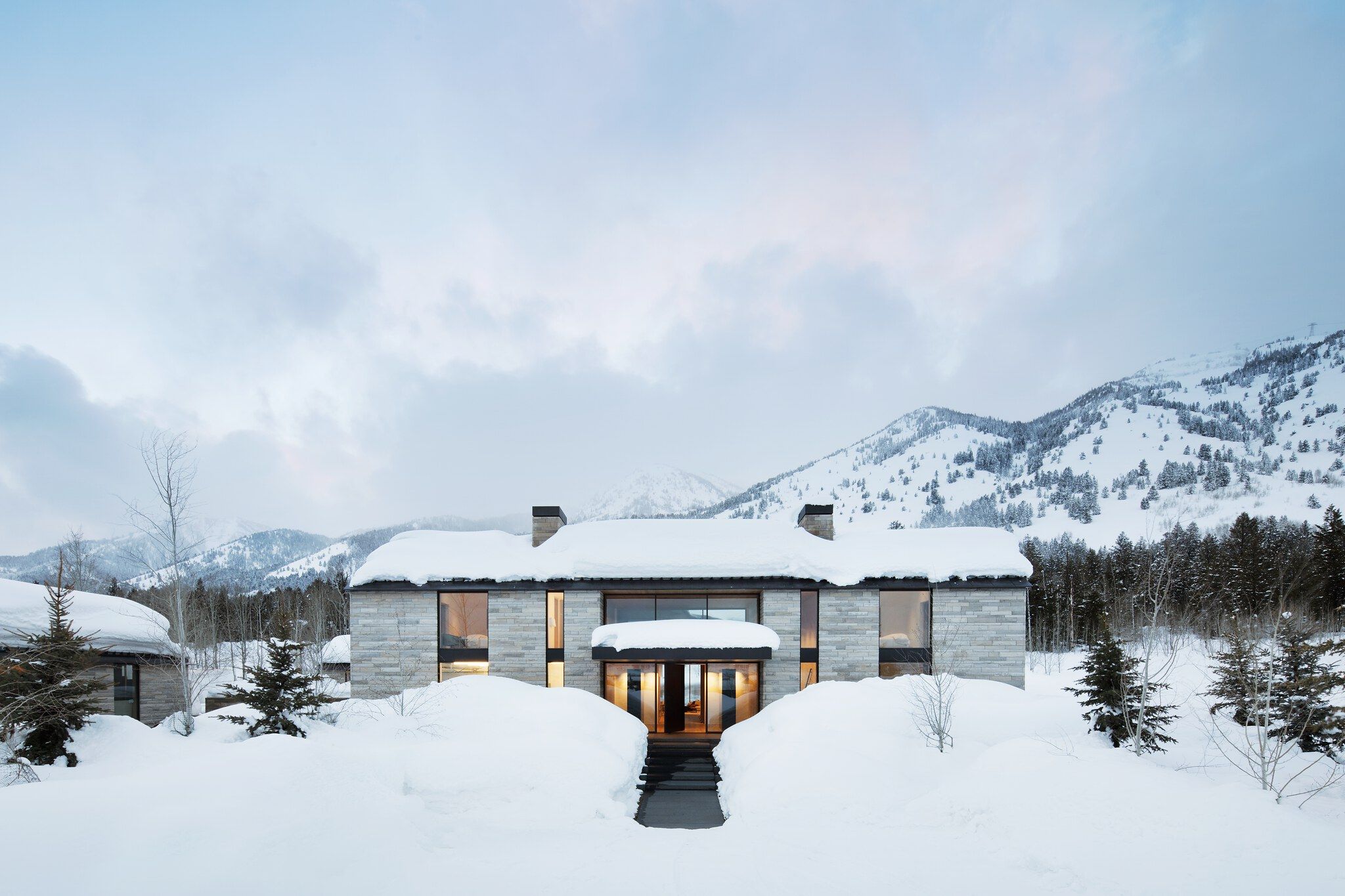 Modern stone house with illuminated windows and a cleared entrance path, nestled in a deep winter mountain landscape. The residence and surrounding ground are covered in heavy snow, with evergreen trees and snow-capped peaks in the background under a cloudy sky.