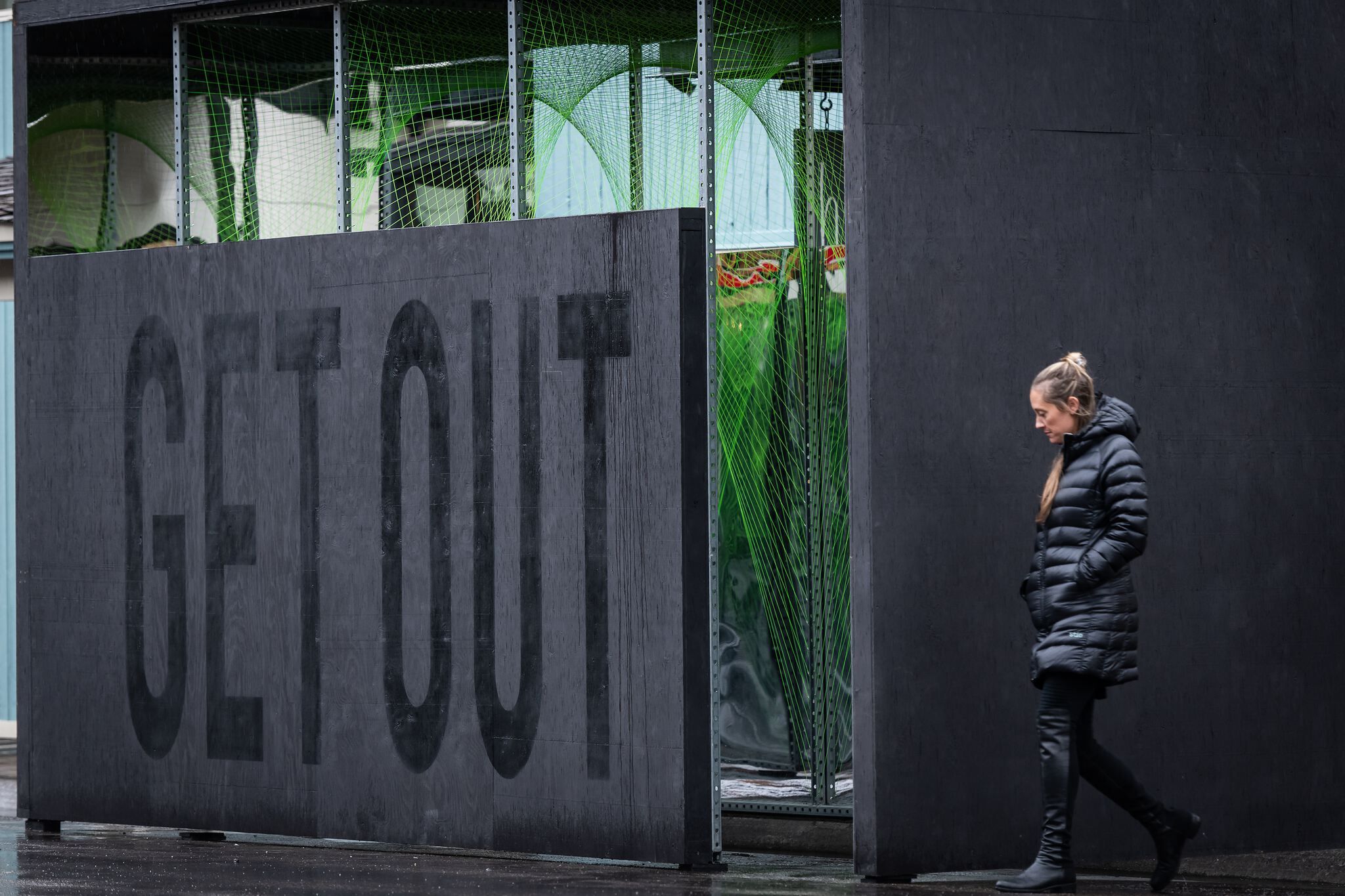 Woman in a black puffer coat and dark leggings, hands in pockets, walks in profile past a tall, dark gray wooden wall with the words GET OUT faintly visible. Behind the wall, a vibrant green netted structure is partially visible. The wet street surface reflects the light.