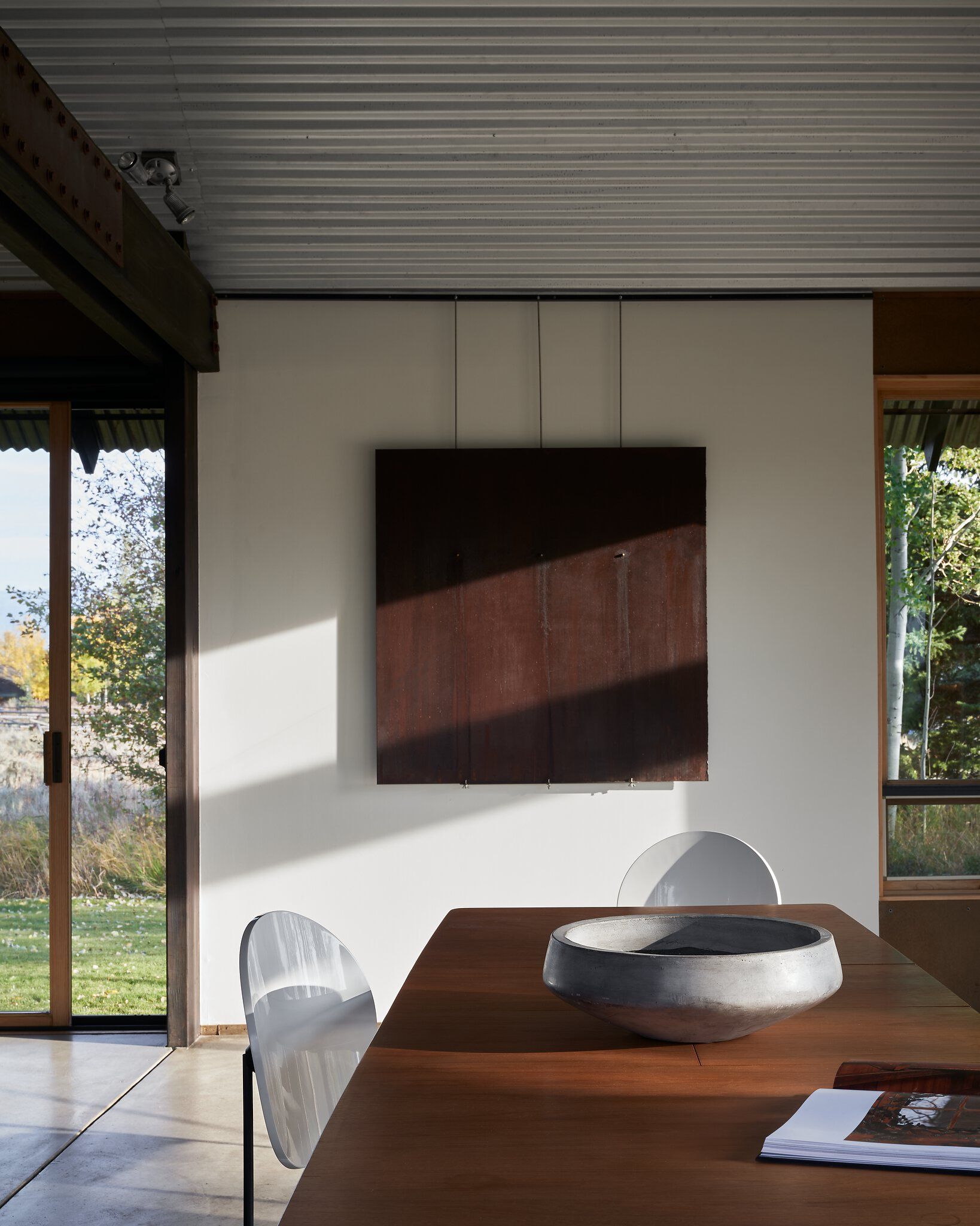 Modern dining area featuring a large wooden table with a concrete bowl and an open book. Sunlight casts diagonal shadows across a white wall displaying a dark, textured metal artwork. Large windows offer views of an autumn landscape with trees and grass, beneath a corrugated metal ceiling and an exposed riveted beam.